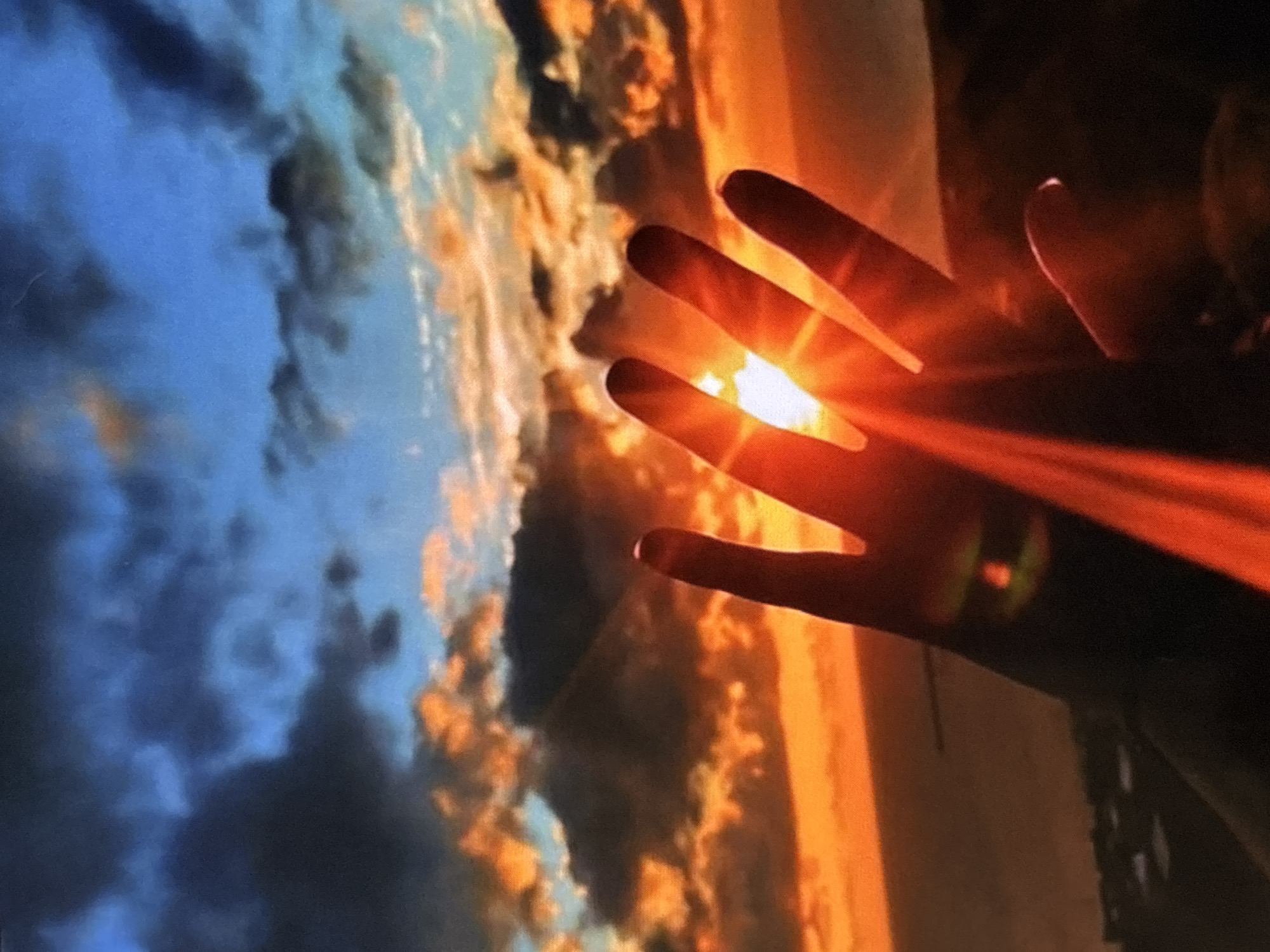 The image shows a hand held up against a sky at sunset or sunrise. The hand is silhouetted by the bright light of the sun, which is partially obscured by the fingers. Red rays of light emanate from between the fingers. The background is a sky with orange and blue clouds.
There is no information in the image to indicate that this image was taken in Belopoptsi, Bulgaria.