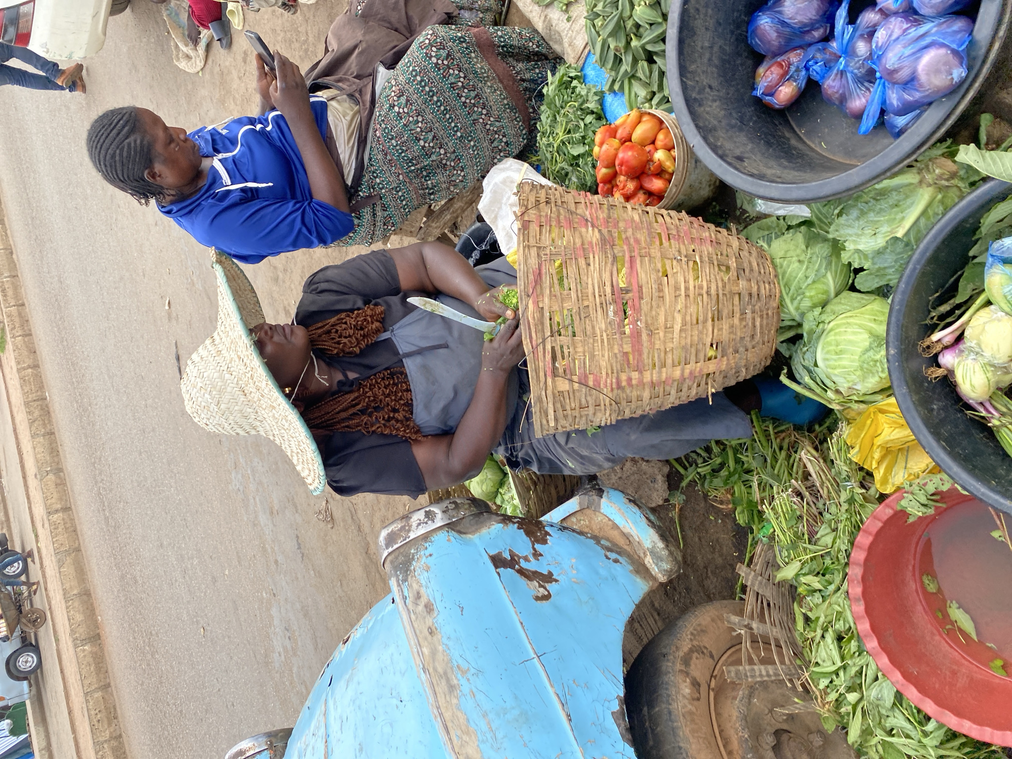 The image depicts a street scene in Bukuru, Nigeria, with a focus on a market setting. Two women are the central subjects. One woman, wearing a blue hooded sweatshirt and with her hair in braids, is seated and looking at a mobile phone. The other woman, wearing a straw hat and a dark top, is kneeling and appears to be preparing vegetables with a knife. She has braided hair and is working near a woven basket.
The scene is filled with various fresh produce. Large black bowls hold onions, tomatoes, and cabbage. Smaller containers and baskets are also visible, filled with more vegetables and leafy greens. A large blue object, possibly part of a vehicle or cart, is prominently featured in the foreground, showing signs of wear and tear. The ground is a mix of dirt and pavement, indicating an outdoor market area. The presence of fresh produce and the interaction between the women suggest a commercial activity, likely a sale or preparation for sale of goods at a market. The lighting suggests it is daytime. No visible text is present in the image.