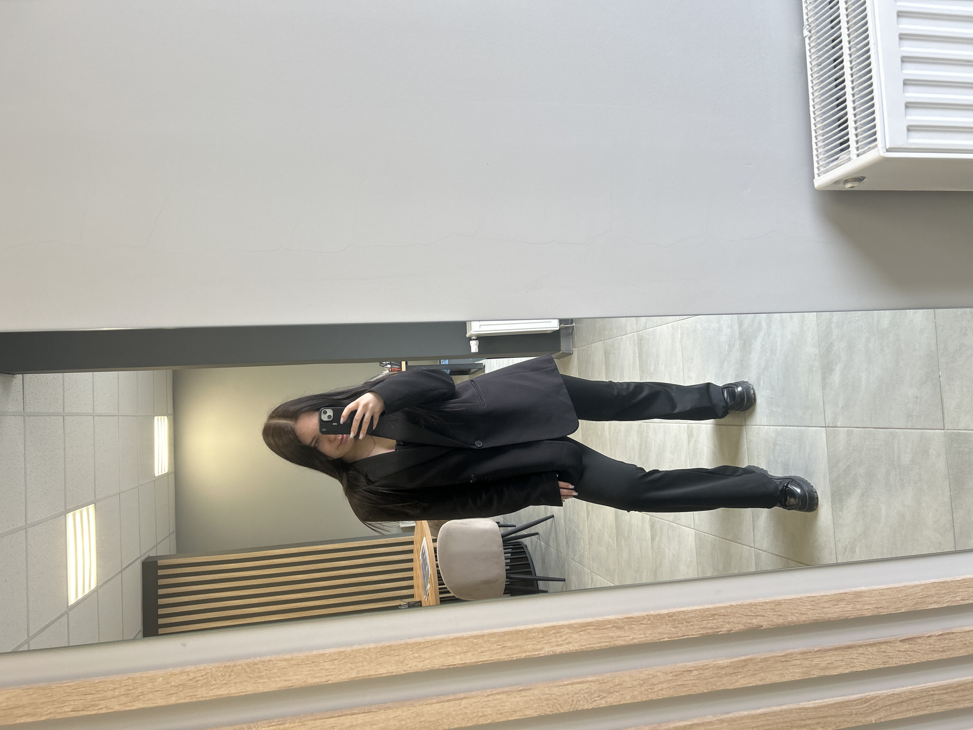 A female individual with long, dark hair stands in an indoor setting, reflected in a full-length mirror. She is wearing a black oversized blazer, a black top, black wide-leg trousers, and black chunky-soled loafer-style shoes. Her right hand holds a black smartphone, seemingly an iPhone, positioned to capture a mirror selfie. Her expression is a slight smile.
The reflected environment behind her includes a light-colored wooden slat decorative partition on the left. Further into the room, a round wooden table with a black base is visible, next to which sits a light-colored, velvet-upholstered chair with black legs. Papers or magazines are on the table. The wall behind the table appears to be a dark grey or green. A doorway or opening to the far right of the reflection reveals a partial view of a desk with various office supplies. The ceiling features white grid tiles with two illuminated rectangular LED panel lights. The floor consists of light grey square tiles.
To the right of the mirror, an actual light grey wall with minor surface imperfections is visible. A white panel radiator is mounted on this wall near the bottom.
The specific location within the city of Izhevsk, Russia, cannot be determined from the visual information provided in the image.