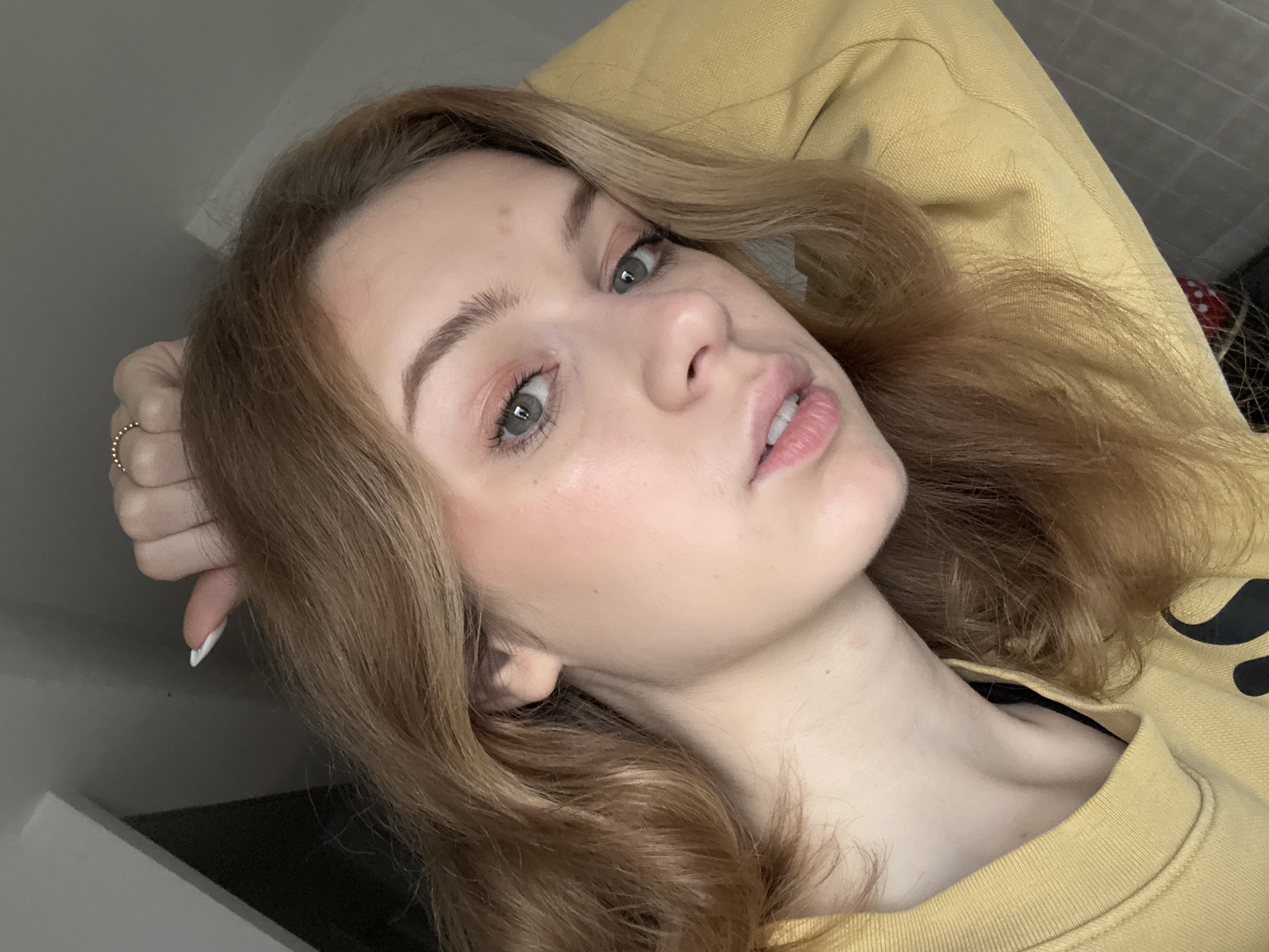 A close-up photograph shows a young woman with light brown hair and fair skin. She is wearing a yellow sweater and a ring on her finger. Her face is tilted, and she is looking towards the camera. The background appears to be a simple room with a gray wall. There is no information to indicate this image was taken in Kragujevac, Serbia, or any other specific location.