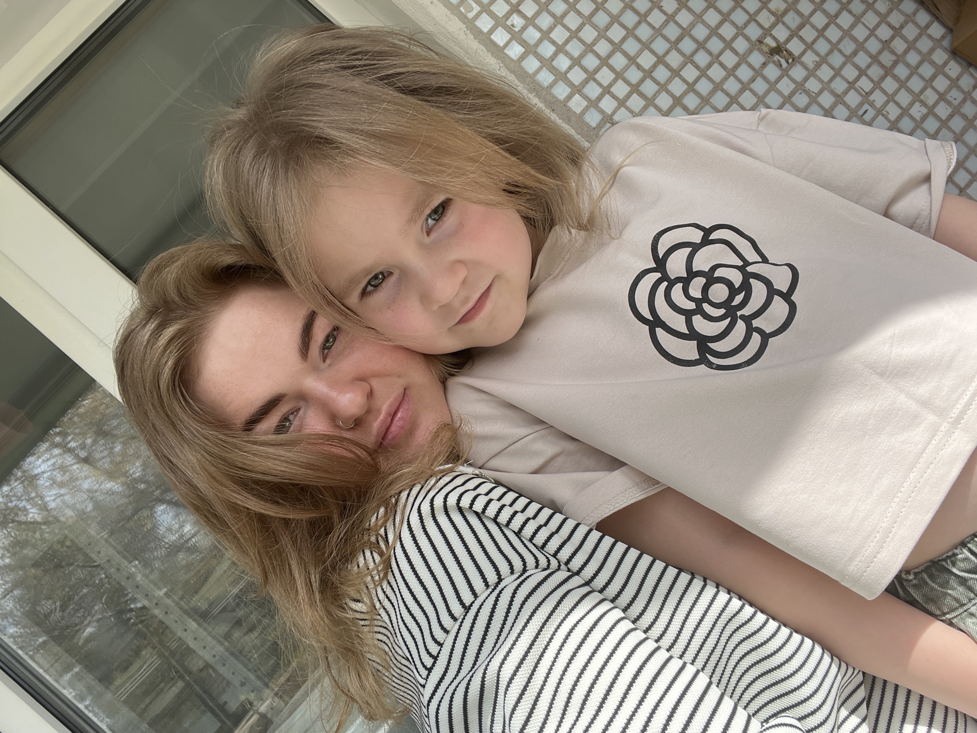 A young woman and child, possibly mother and daughter, are posing close together for a photo. The woman has shoulder-length blonde hair, fair skin, and a nose ring. She wears a black and white striped top. The child has blonde hair, fair skin, and wears a light beige t-shirt with a black flower design. They appear to be indoors, as indicated by the presence of a window in the background reflecting outdoor scenery, possibly trees. The location of the photo is Chaikovsky, Russia.