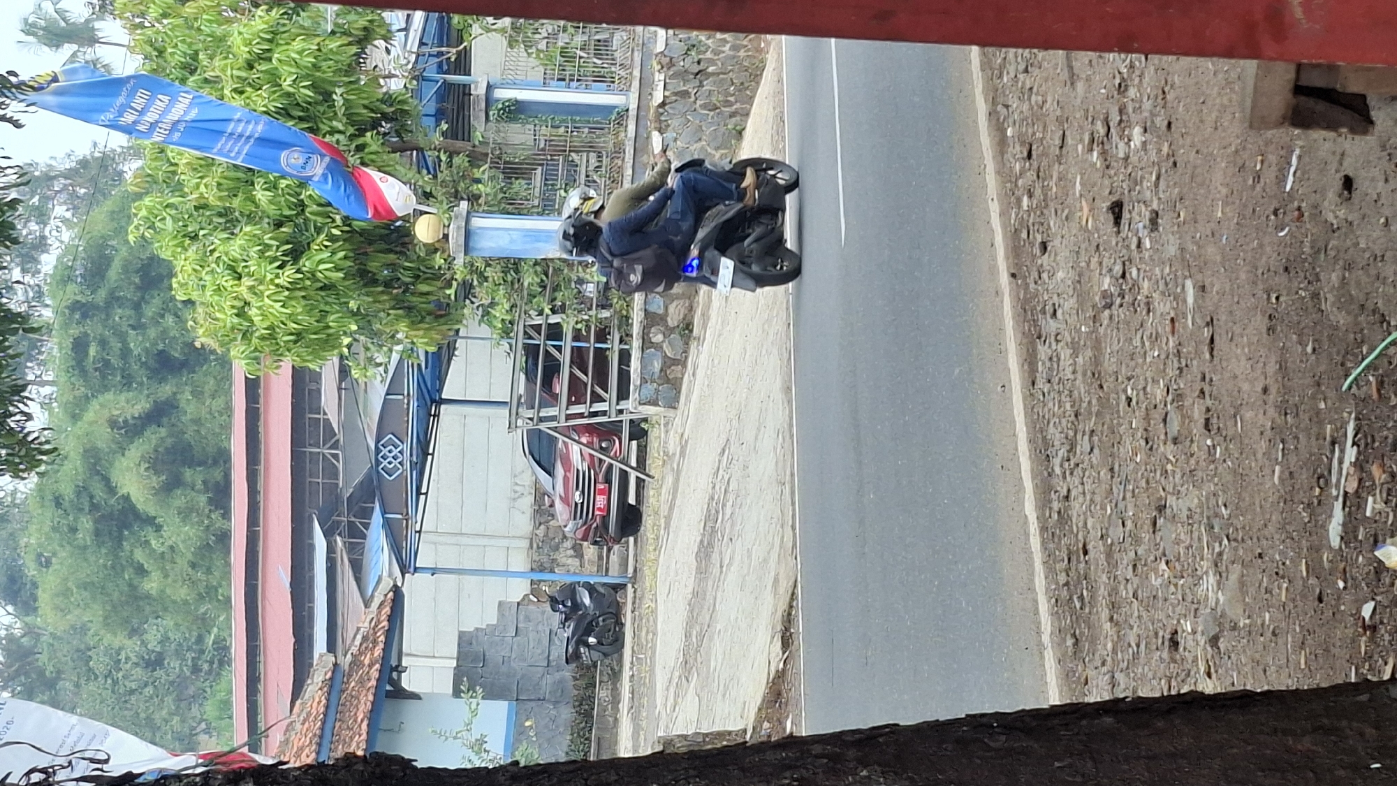 In the city of Cilaku, Indonesia, a red sedan is parked in front of a building with a blue awning. To the left of the sedan, a black motorcycle is parked. In the foreground, two people are riding on a black motorcycle. The rider in front is wearing a blue jacket and a helmet. The person behind them is wearing a dark jacket and a helmet. A blue light is visible on the back of the motorcycle. The scene appears to be on a street or road, with trees and vegetation in the background. A large blue flag with white and red stripes is hanging from a pole on the right side of the image.