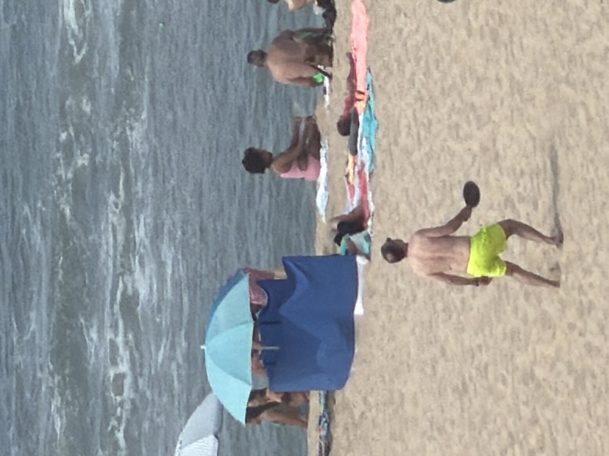 The image depicts a beach scene on a sunny day, with the ocean in the background. In the foreground, a man wearing neon yellow swim shorts is seen from the back, holding a paddle in his right hand, appearing to be playing a paddle game on the sandy beach. Further back, several people are relaxing on towels. A blue beach shelter is set up, with a light blue umbrella above it. To the left, another beach umbrella is partially visible. In the middle ground, a woman in a pink swimsuit is sitting, and behind her, a man is seated with his back to the camera. The environment is a typical beach setting with sand and the ocean. This scene is consistent with a day at the beach in Espinho, Portugal, known for its sandy shores and recreational activities.