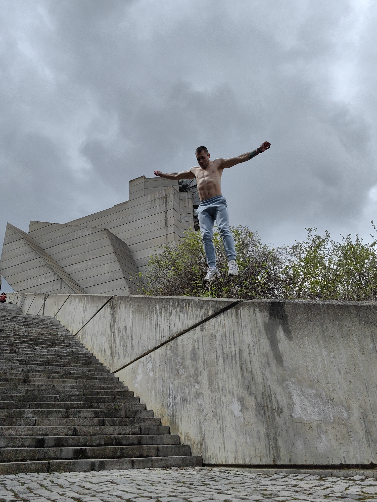 A shirtless man in blue pants jumps in front of the Monument to 1300 Years of Bulgaria, located in Shumen, Bulgaria. The man is mid-air with arms outstretched. He wears white athletic shoes. He is jumping off of a concrete retaining wall that runs along a set of stairs leading up to the monument. The monument itself is a large, concrete structure composed of geometric shapes. The sky is overcast. Green vegetation is visible behind the retaining wall. A person in a red jacket is on the stairs. The ground is paved with cobblestones.