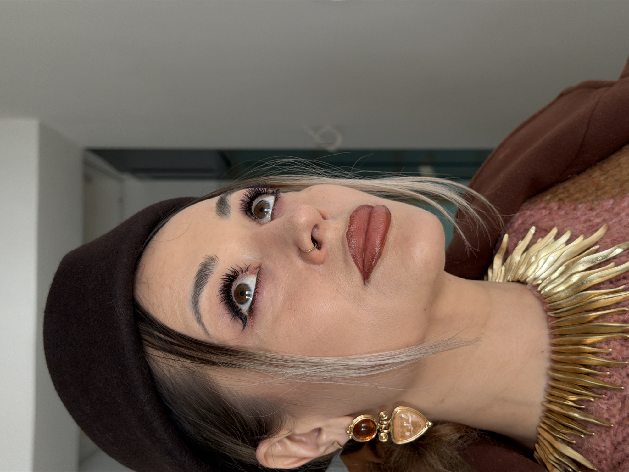 A woman is seen from a low angle, her head tilted back. She wears a dark brown beret and has light brown hair with blonde streaks framing her face. Her makeup includes prominent eyelashes, brown lipstick, and subtle red eyeliner. She also has a small gold nose ring. She is wearing a gold, spiked collar necklace and amber earrings. Her clothing appears to be a brown jacket and a pink sweater. The background is a neutral gray. There is no information available to confidently place the location in the city of Krasnoyarsk, Russia.