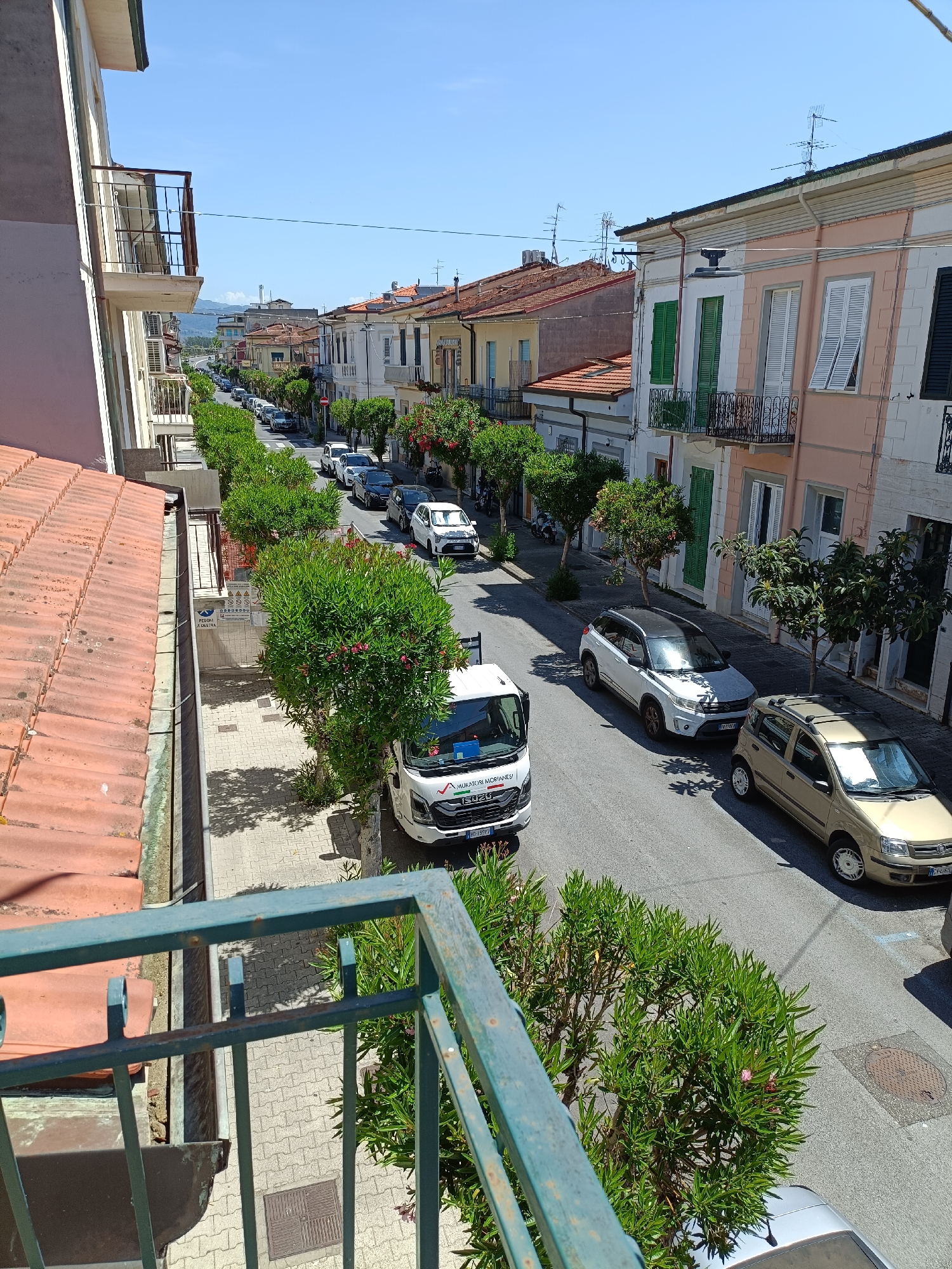 The image depicts a sun-drenched street scene in Viareggio, Italy, likely taken from a balcony. The architecture features buildings with colorful facades, tiled roofs, and balconies adorned with railings, characteristic of Italian coastal towns. Green trees with small pink flowers line the street, casting dappled shadows on the paved sidewalk and asphalt road.
Several vehicles are parked and driving along the street. These include a white Isuzu truck with visible text on its side that appears to be "MURATORI MORANELLI" and "ISUZU," a white SUV, a light-colored hatchback, and a silver car. The presence of these cars suggests a normal day with local traffic and perhaps some residents or visitors.
The time of day appears to be late morning or early afternoon, indicated by the bright sunlight and distinct shadows. The weather seems to be clear and warm, with a bright blue sky dominating the upper portion of the image. The overall mood is tranquil and everyday, capturing a slice of life in a Mediterranean town. A few pieces of visible text, primarily on the truck, offer clues to local businesses. The scene is one of everyday activity on a pleasant day in a residential or mixed-use street.