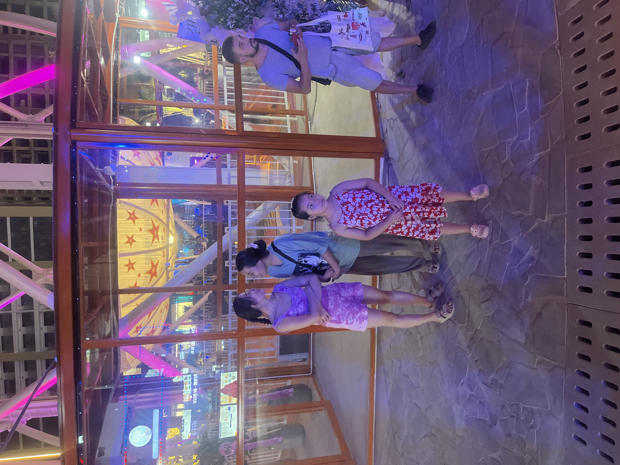 This image captures a scene at the Sun Wheel in Đà Nẵng, Vietnam, likely at dusk or night, given the bright artificial lighting. The foreground features a paved ground with a drainage grate. Three women and one man are visible. Two women, appearing to be of Asian descent, are standing in the middle left, facing each other. One is wearing a purple tank top and patterned shorts, with her arms crossed. The other is in a green t-shirt and dark pants, with a crossbody bag. To their right, a young girl with dark hair is wearing a red and white floral dress. On the far right, a man with short dark hair and a beard is wearing a light blue t-shirt and light-colored shorts. He has a dark strap across his chest and is holding a white tote bag with graphics and a red object in his right hand. Behind the individuals, a glass-enclosed structure with orange framing is visible, revealing the colorful lights and support beams of the Sun Wheel in the background. The environment suggests an amusement park or a recreational area with vibrant illumination.