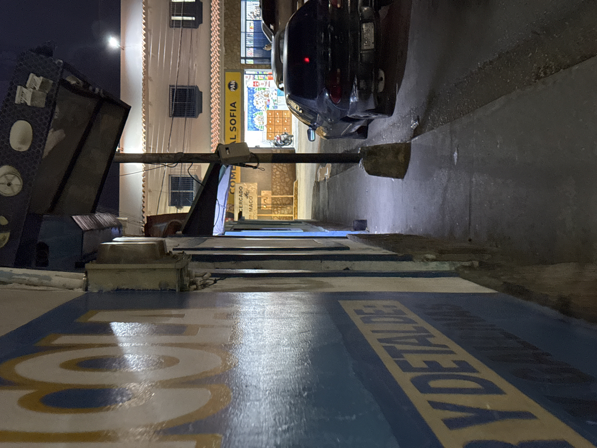 The image depicts a street scene at night in Santa Ana, Venezuela. On the left, a blue and white wall features large yellow lettering that appears to be part of a business sign, with visible words like "Y DETALDE". To the right, a sidewalk runs alongside the wall, leading towards a building with a tiled roof. This building houses a store with "AL SOFIA" and "PAN" visible on its yellow awning. Inside the store, shelves and a colorful mural are discernible. A black sedan is parked on the street in front of the store, and a motorcycle is visible just outside the store entrance. The street is paved, and a concrete pole with electrical conduits stands near the sidewalk. The overall environment suggests a commercial area within a town.