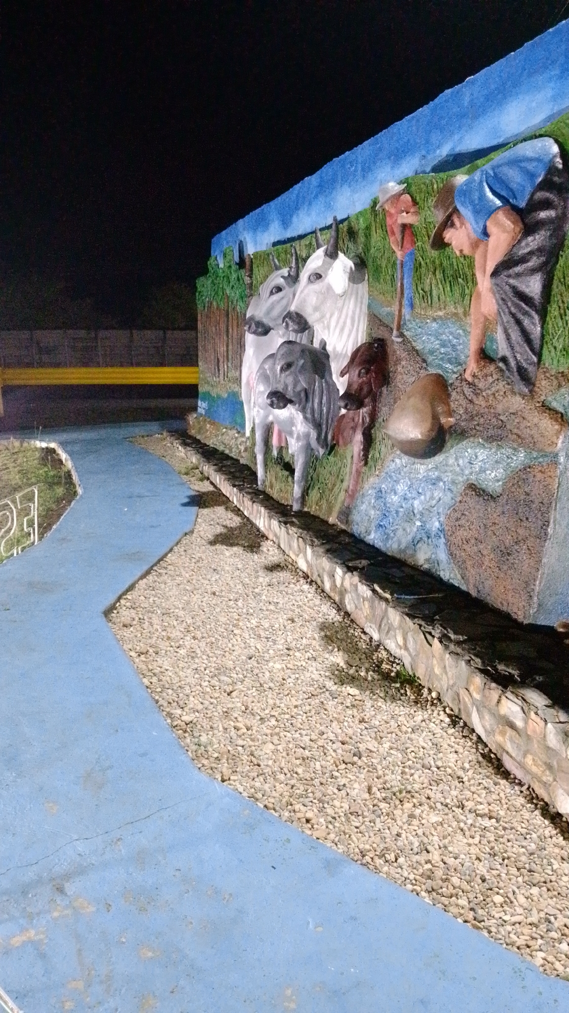 A mural in Tinaquillo, Venezuela depicts three cattle (two white and one gray) and a brown calf standing by a stream. The mural also depicts two men: one bent over the stream, and another holding a shovel. The mural is built on a stone foundation. The foreground includes a blue-painted walkway and an area of light-colored pebbles. In the background, there is a fence made of yellow boards. The sky above is dark. The image appears to have been taken at night.