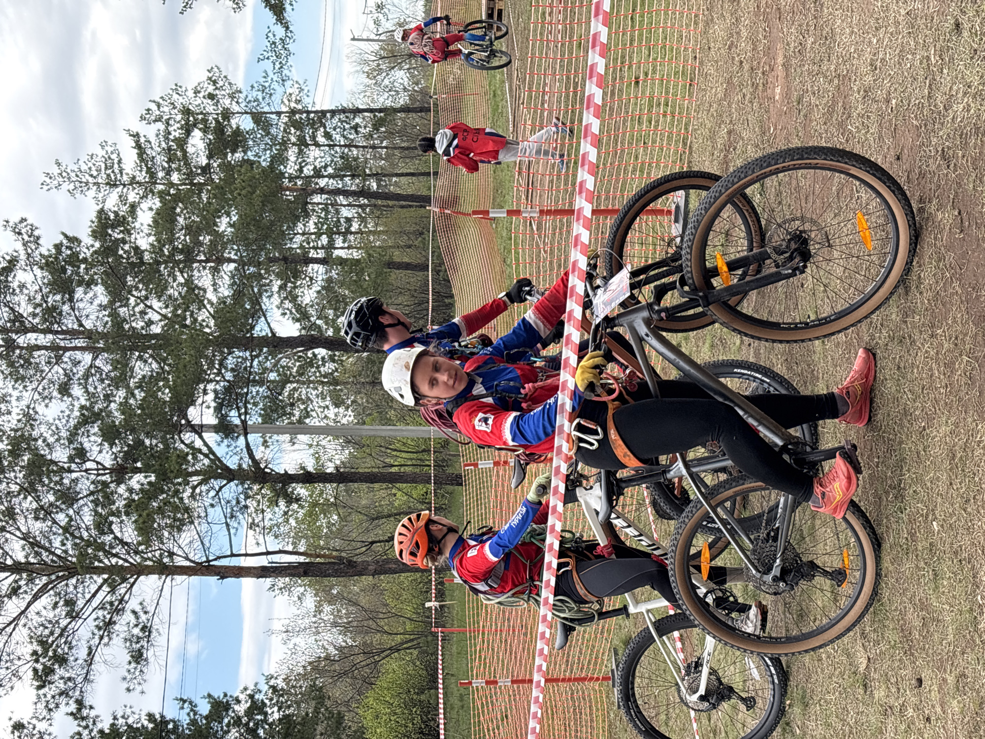 The image shows three individuals preparing for a mountain biking competition, likely in a natural setting within Orenburg, Russia. They are wearing helmets and cycling gear, positioned behind a red and white striped barrier, suggestive of a designated starting area or a restricted zone. They are securing themselves onto their bikes with what appear to be safety ropes. The background includes tall trees and a grassy area, hinting at a park or forest environment on a partly cloudy day. Other participants and bikes are visible further down the path, indicating the presence of a broader competition event.