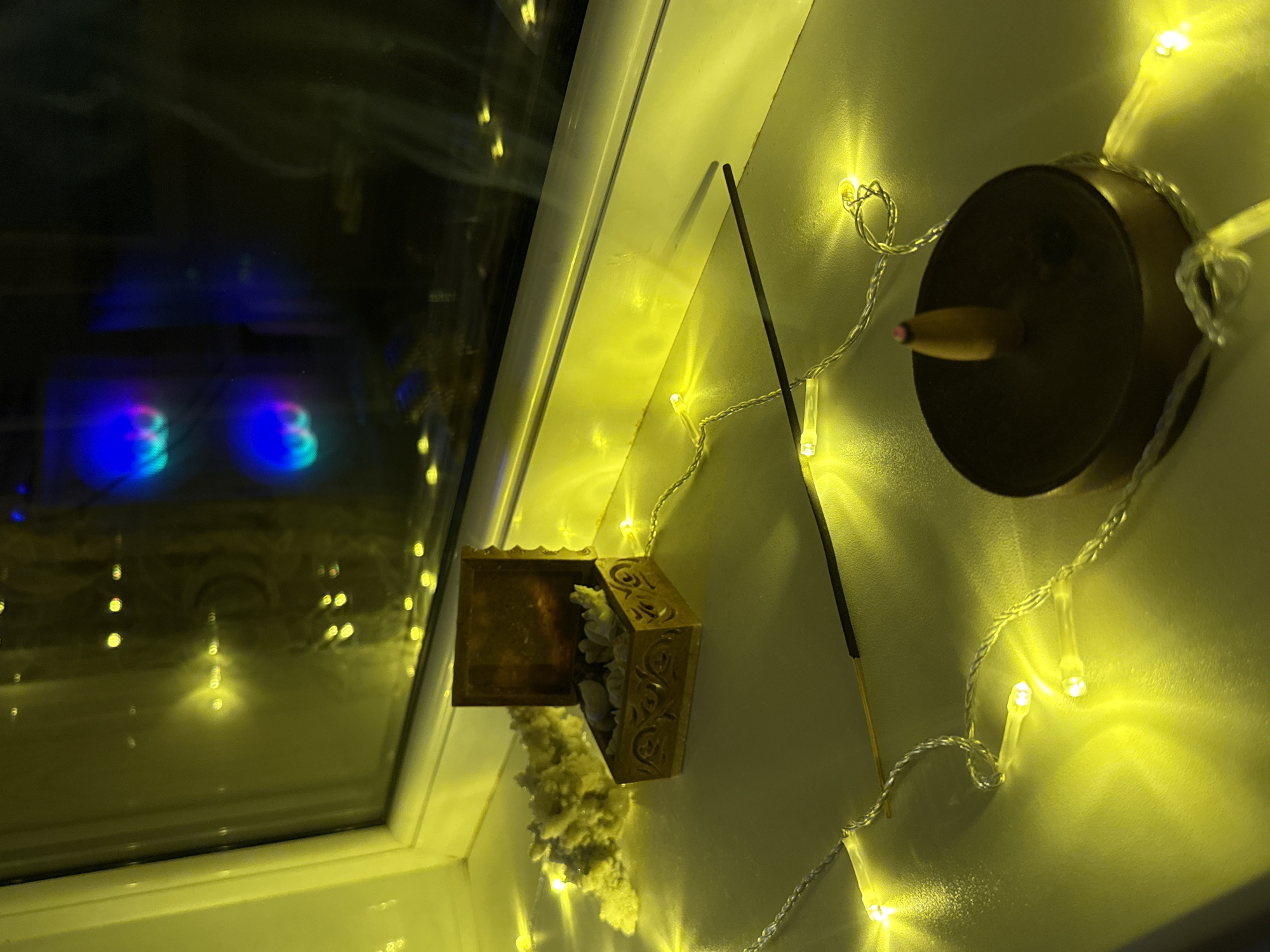 This image, captured in Naberezhnye Chelny, Russia, depicts an indoor scene focused on a white windowsill. The upper half of the frame is dominated by a window with a white frame. The window glass reflects multiple internal yellow light sources, originating from a string of LED fairy lights, and distinctly shows two internally illuminated, multi-colored (blue and pink) circular forms, consistent with computer cooling fans, suggesting a device within the room. The exterior view through the window is dark and indistinct, with faint, distant light sources.
A string of warm yellow LED fairy lights with clear wiring is arranged loosely across the windowsill, serving as the primary illumination for the objects.
On the left side of the windowsill, an open, ornate, rectangular wooden box, featuring carved patterns, contains several small, irregularly shaped, light-colored objects, possibly pebbles or shells. Behind this box, and resting against the window frame, is a white, textured, branch-like object, resembling coral or a dried botanical element.
A thin, dark brown or black incense stick lies horizontally across the central portion of the windowsill, partially resting on the fairy lights. Its tip, pointed towards the left, appears unburnt or golden.
On the right side of the windowsill, a dark, circular, flat-topped incense burner is present. A small, light-colored, conical element protrudes from its center, with the very tip displaying a subtle reddish glow, indicating it may be lit or recently extinguished. No human subjects or active interactions are visible within the frame.