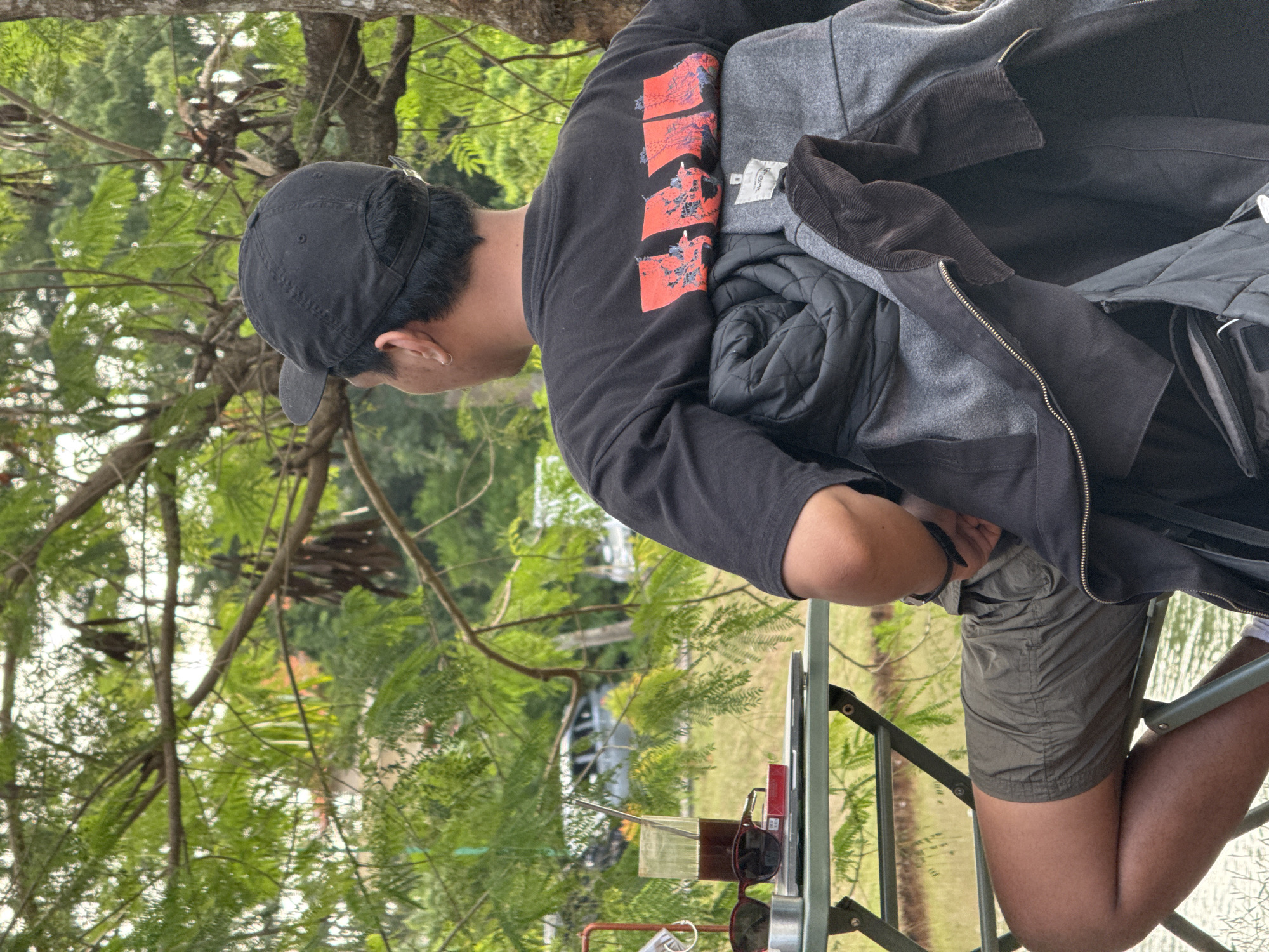 A man with short black hair and a black cap is sitting outside near a body of water. He is wearing a black shirt with red graphic designs on the sleeve and grey shorts. He has multiple jackets draped over his shoulders, including a grey jacket with a black corduroy collar. He is at a table with sunglasses, a lighter, and other items. Green foliage and trees are visible in the background. Based on the prompt, the location is Kuta Selatan, Indonesia.