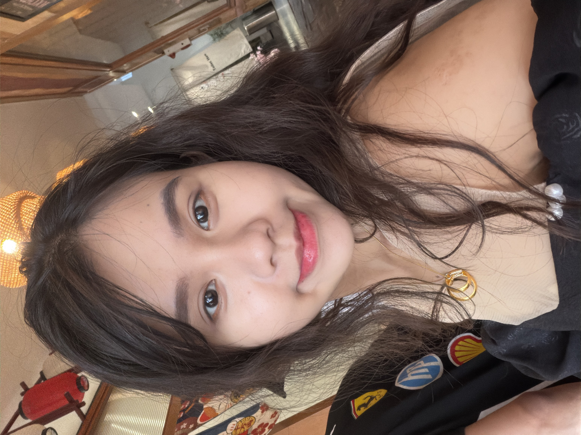 A young woman is captured in a close-up, slightly tilted portrait, possibly a selfie, within an indoor setting. She has long, dark, slightly wavy hair that frames her face and shoulders. Her eyes are dark, and she wears pink lipstick, sporting a gentle, pleasant smile. A small mole is visible under her left eye.
She is wearing a dark-colored top, possibly black, with several embroidered patches on the right shoulder area. Visible patches include the yellow and red Shell logo, a yellow shield with the black Ferrari prancing horse, and a blue shield-like emblem. A lighter, flesh-toned garment is worn underneath, visible near her neck and shoulder. A delicate gold-colored necklace with two interlocked rings as a pendant rests on her chest. A pearl earring is visible on her left ear.
The background is softly blurred but shows warm tones and various elements. To the left, a woven rattan light fixture emits a soft glow. Below it, a red, lantern-like decorative object is partially visible. A patterned fabric with floral or geometric motifs in red, blue, and white is also seen in the lower left. In the upper right and center background, reflections on a surface reveal parts of an interior, with faint, blurry text that appears to read "Toilet Here". The overall decor, including the rattan lamp and red accent, suggests a cafe or restaurant environment, consistent with a location in Quy Nhon Ward, Vietnam. The lighting is soft and even, indicating an indoor setting. The woman's expression is calm and relaxed.
