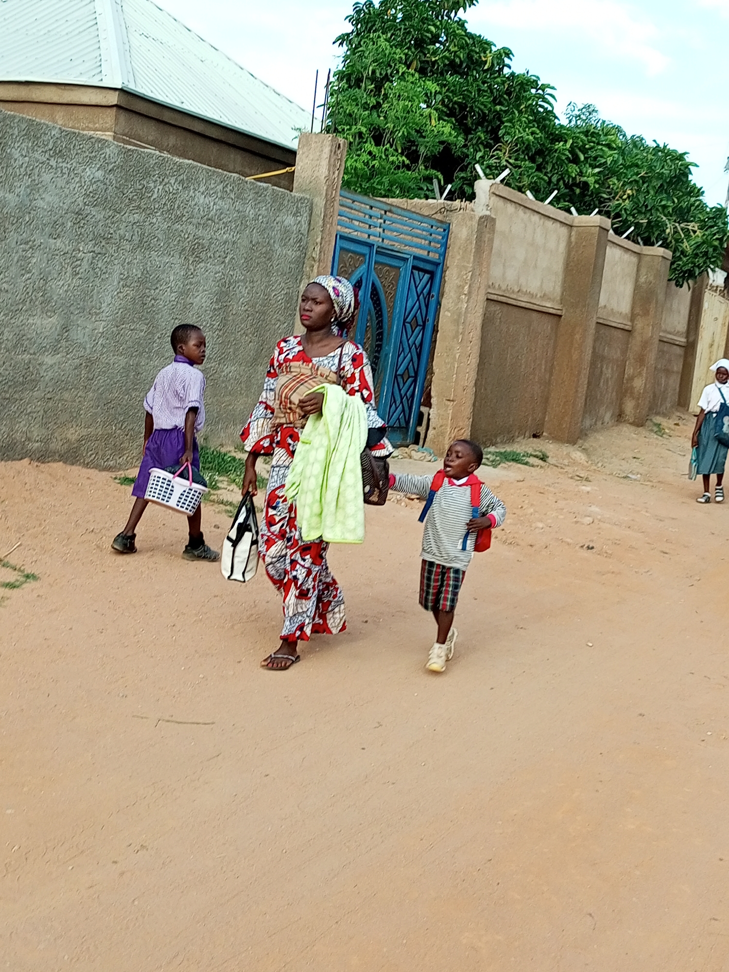 In an outdoor scene, a group of people walks on a dirt path beside a wall and a blue gate. A woman in a brightly patterned dress and headscarf walks forward, carrying a light green cloth and a patterned tote bag. She is accompanied by two young children. A boy wearing a striped shirt, plaid shorts, and a red backpack walks towards the right, his mouth open as if speaking or shouting. Another boy in a purple uniform and shorts walks towards the left, carrying a white rectangular basket with pink handles. In the background, a woman in a white shirt and blue skirt walks further down the path. The environment consists of a rough-textured wall, some greenery, and a cloudy sky.
