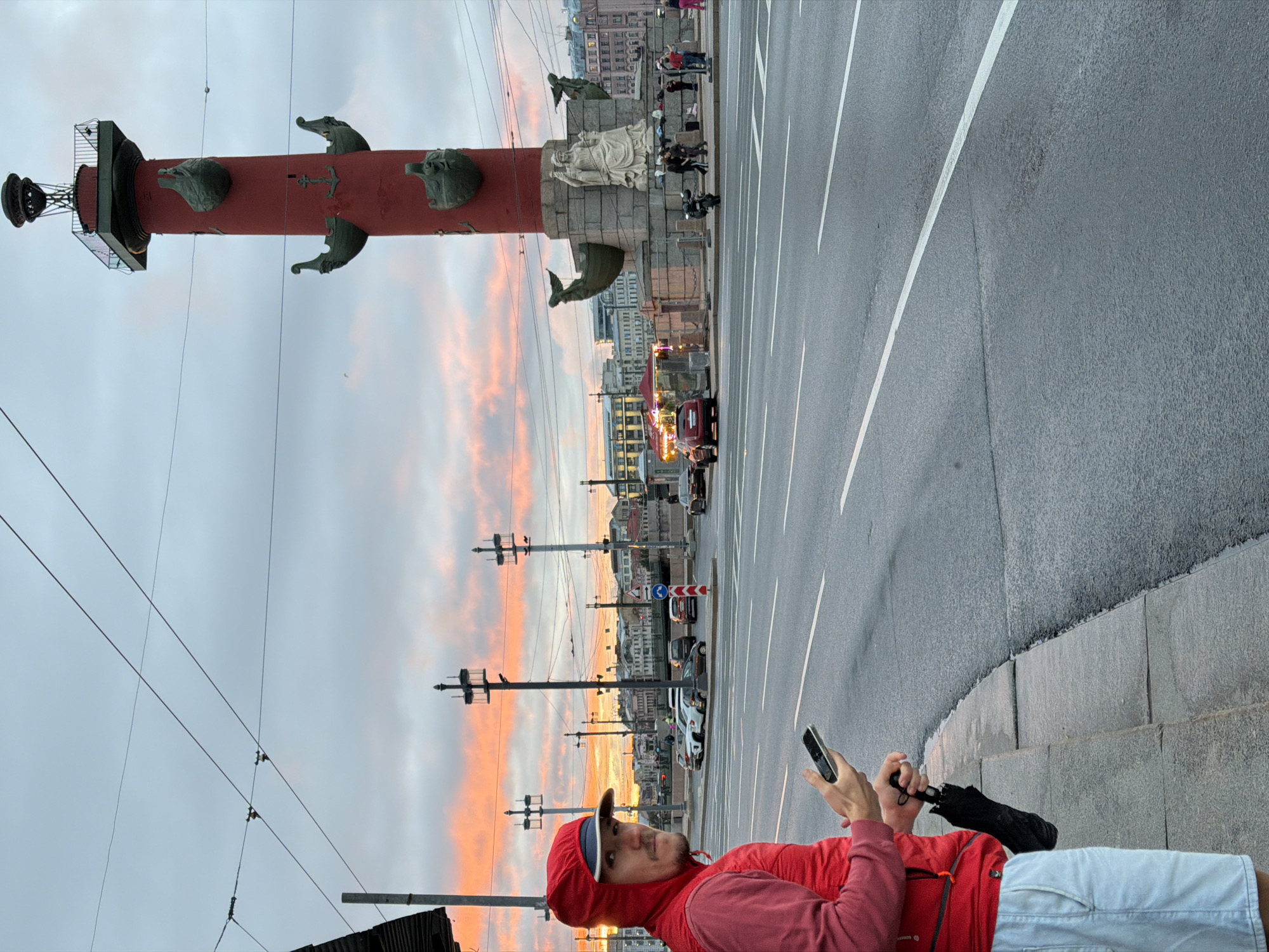 An outdoor, medium-shot photograph taken at dusk captures a man in the foreground, standing on a sidewalk next to a street with moderate traffic. The man, wearing a red hooded jacket and light blue denim shorts, is looking towards the camera and holding a smartphone in his right hand and a folded black umbrella in his left. He appears to be waiting.
Behind the man, a wide asphalt road stretches into the distance, lined with streetlights and utility poles. Across the street, a prominent red Rostral Column, a landmark in Saint Petersburg, Russia, stands tall against a sky painted with dramatic sunset colors. The column is adorned with sculptures of ships and anchors. A statue of a seated female figure is visible at the base of the column.
In the background, buildings of various architectural styles line the cityscape, illuminated by the fading light. Several cars are driving on the road, and a few people are seen near the column. The overall atmosphere is that of a city winding down at the end of the day.