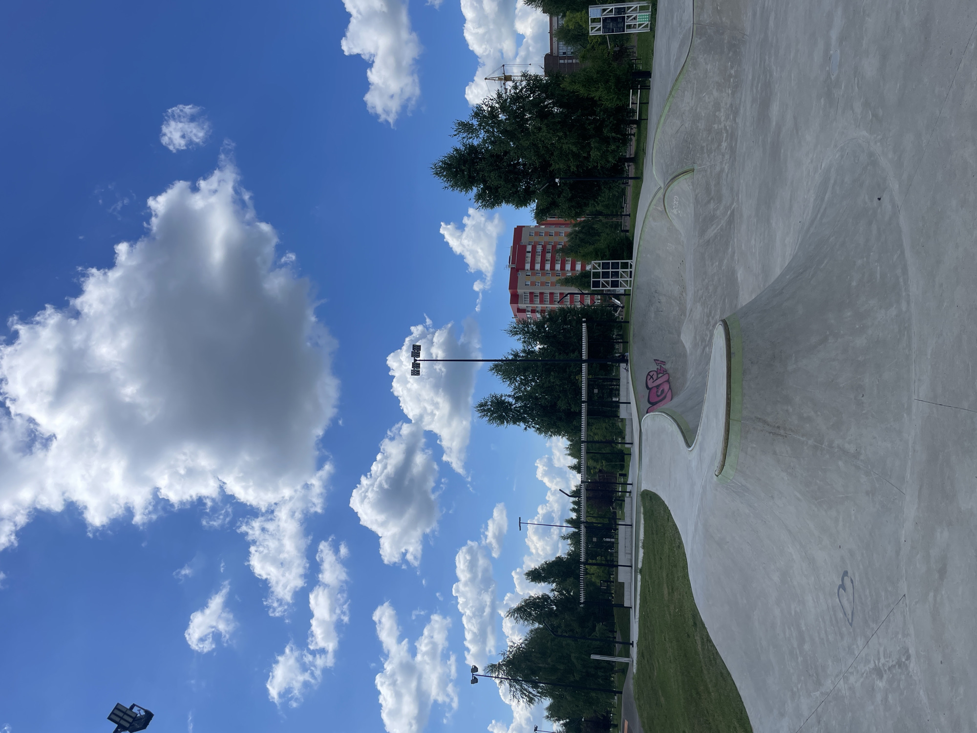 The image shows a concrete skatepark with various ramps and bowls, located in Laishevo, Russia. The skatepark is situated in a park-like setting with green grass and numerous trees surrounding it. In the background, there are residential buildings, one of which is a tall, red and yellow apartment complex. A construction crane is also visible in the distance, suggesting ongoing development in the area. The sky is a vibrant blue with fluffy white clouds, indicating fair weather. There are no people or animals visible in the frame, and no skateboards or other sports equipment are present. The scene is illuminated by daylight, with prominent stadium lights mounted on poles throughout the park. The concrete surfaces of the skatepark show some signs of wear and graffiti.