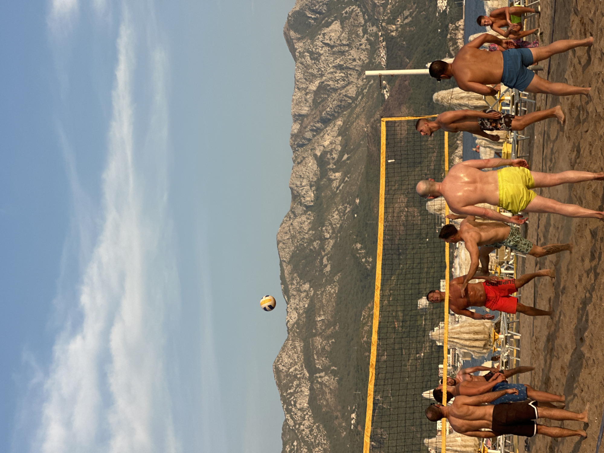In Boreti, Montenegro, a group of men are playing beach volleyball on a sandy court, with a net strung across the middle. The sky is a clear blue with scattered white clouds. In the background, a range of rugged, mountainous terrain stretches across the horizon, with lush green vegetation covering the lower slopes. The upper parts of the mountains are rocky and barren. Some beach umbrellas and lounge chairs are visible behind the volleyball court, suggesting a resort or public beach area. The sun appears to be setting or is in the late afternoon, casting a warm light on the scene.