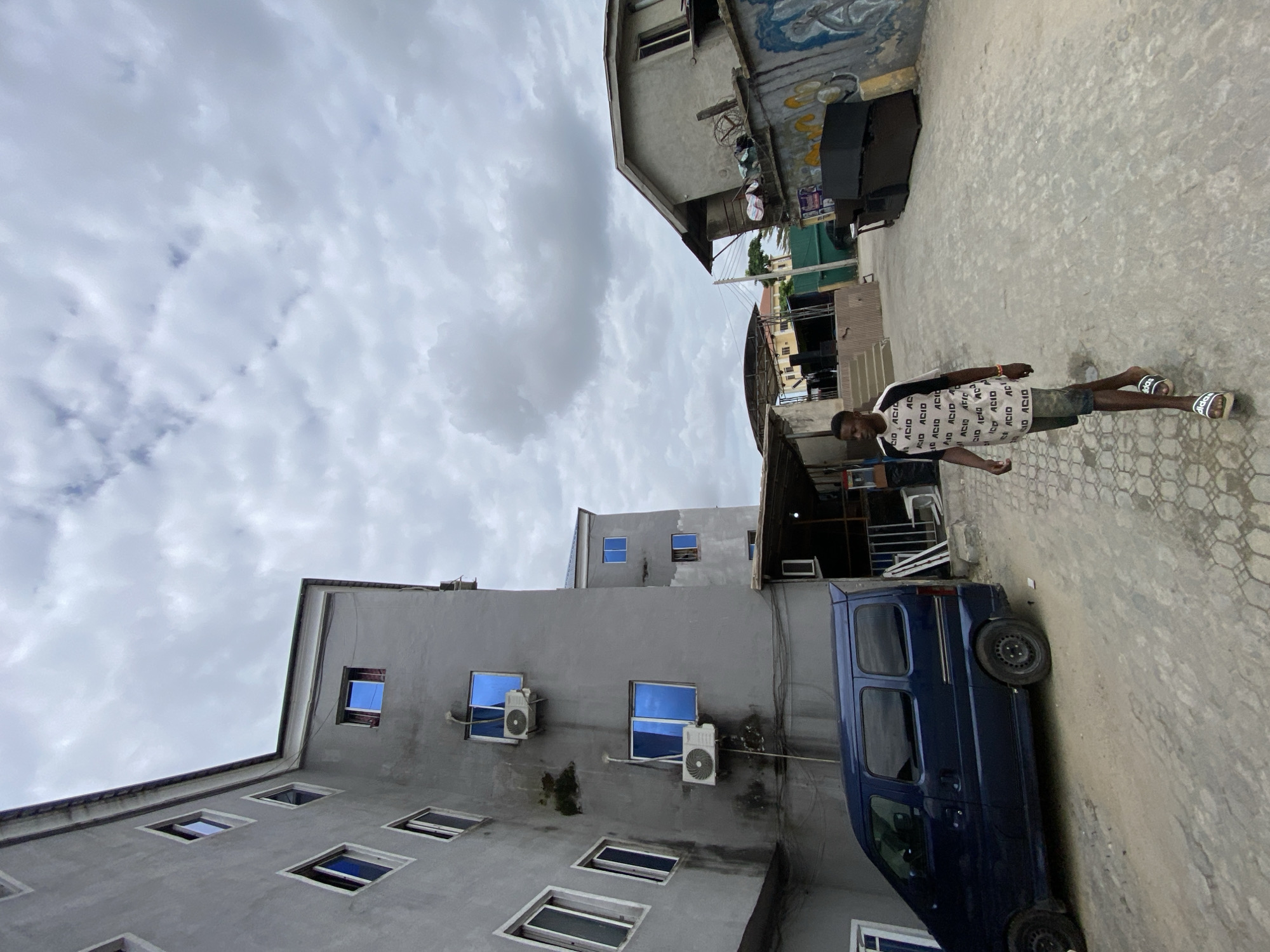 The image depicts a scene in what appears to be Ado, Nigeria. A young man is standing in a narrow alley or street. He is wearing a patterned t-shirt and shorts, with sandals.
The environment consists of buildings on either side of the alley. One building is a tall, gray structure with several windows and air conditioning units. A blue van is parked adjacent to the building. The building on the other side is older and shows signs of wear, with graffiti and other elements present. The sky is overcast with clouds.