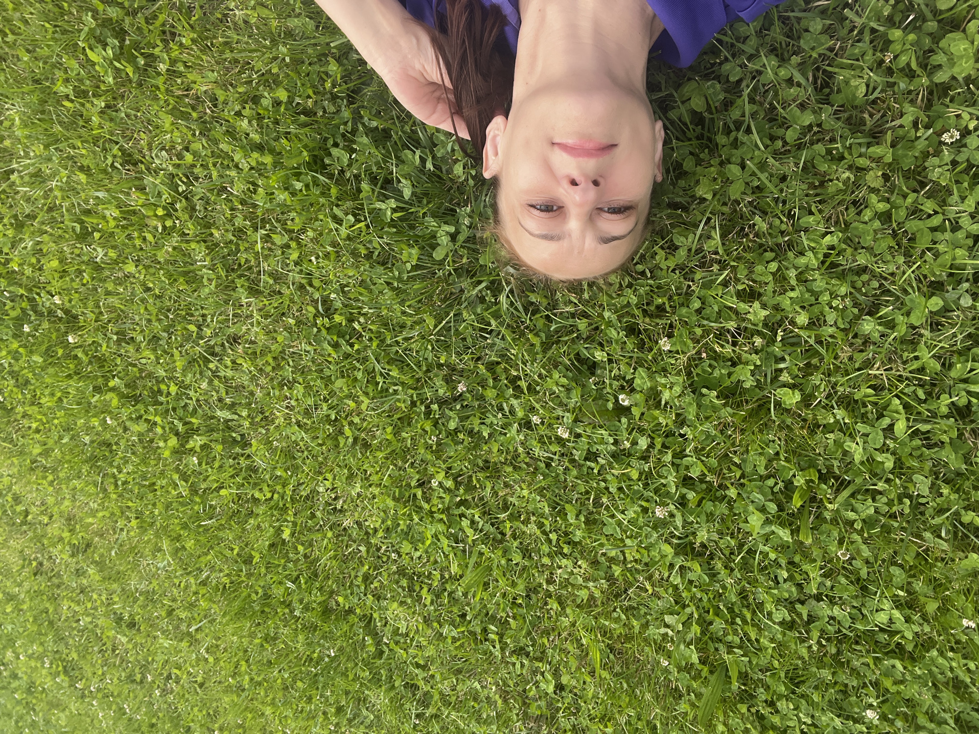 A woman is lying on her back in a grassy field, with her head resting on the grass. The woman is wearing a purple shirt. The grass is green and there are small white flowers scattered throughout. The image is taken from above, looking down at the woman. The location is unknown.