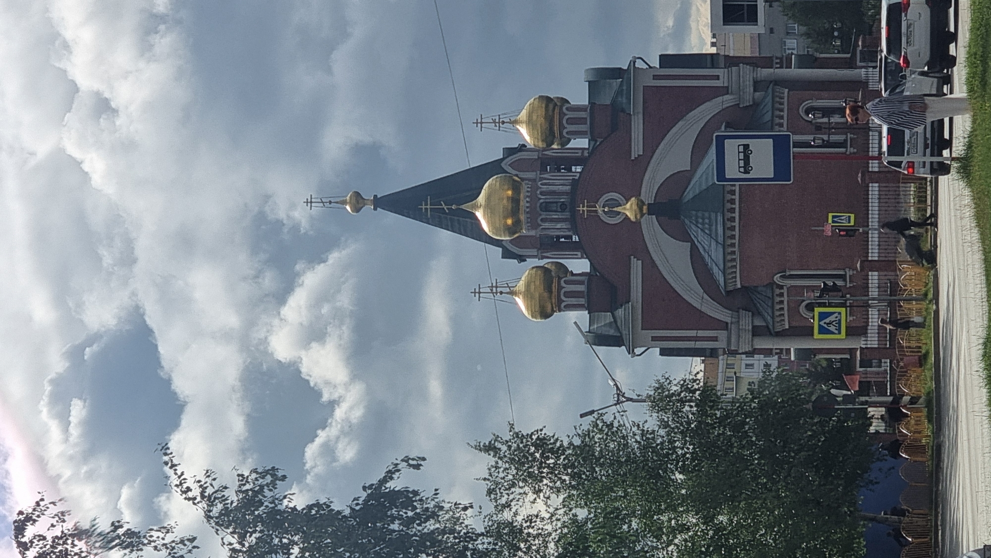 The image shows the Resurrection Cathedral in Megion, Russia, on a cloudy day. The cathedral is a red brick building with multiple golden domes topped with crosses. In the foreground, there is a street with a pedestrian crossing, a bus stop sign, traffic lights, and several cars. People are visible on the sidewalk and crossing the street. To the left of the cathedral, a tree with green foliage is prominent. The sky is filled with dramatic, dark clouds, with some patches of blue sky peeking through.