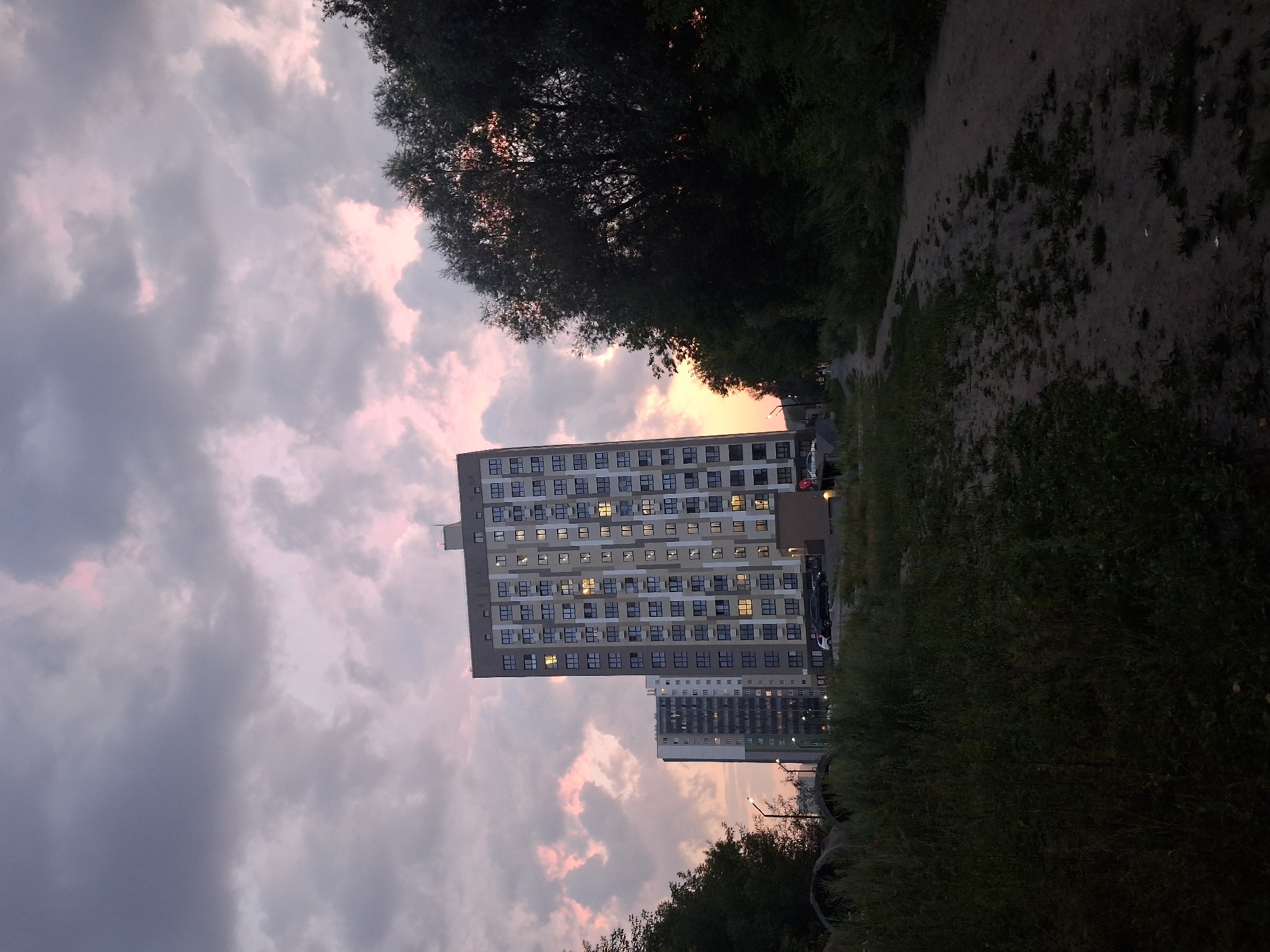 This is a low-angle, full shot of a residential building in Novosibirsk, Russia, at dusk. The building, a modern, multi-story structure with numerous windows, dominates the center of the frame. To the left of it, another, slightly taller building is partially visible. A dirt path or unpaved road curves through the foreground, bordered by overgrown grass and vegetation. Dark trees line the right side of the frame, partially obscuring the sunset. The sky above is filled with dramatic, heavy clouds tinged with pink and orange from the setting sun. There are no visible people, and the scene is still and quiet.