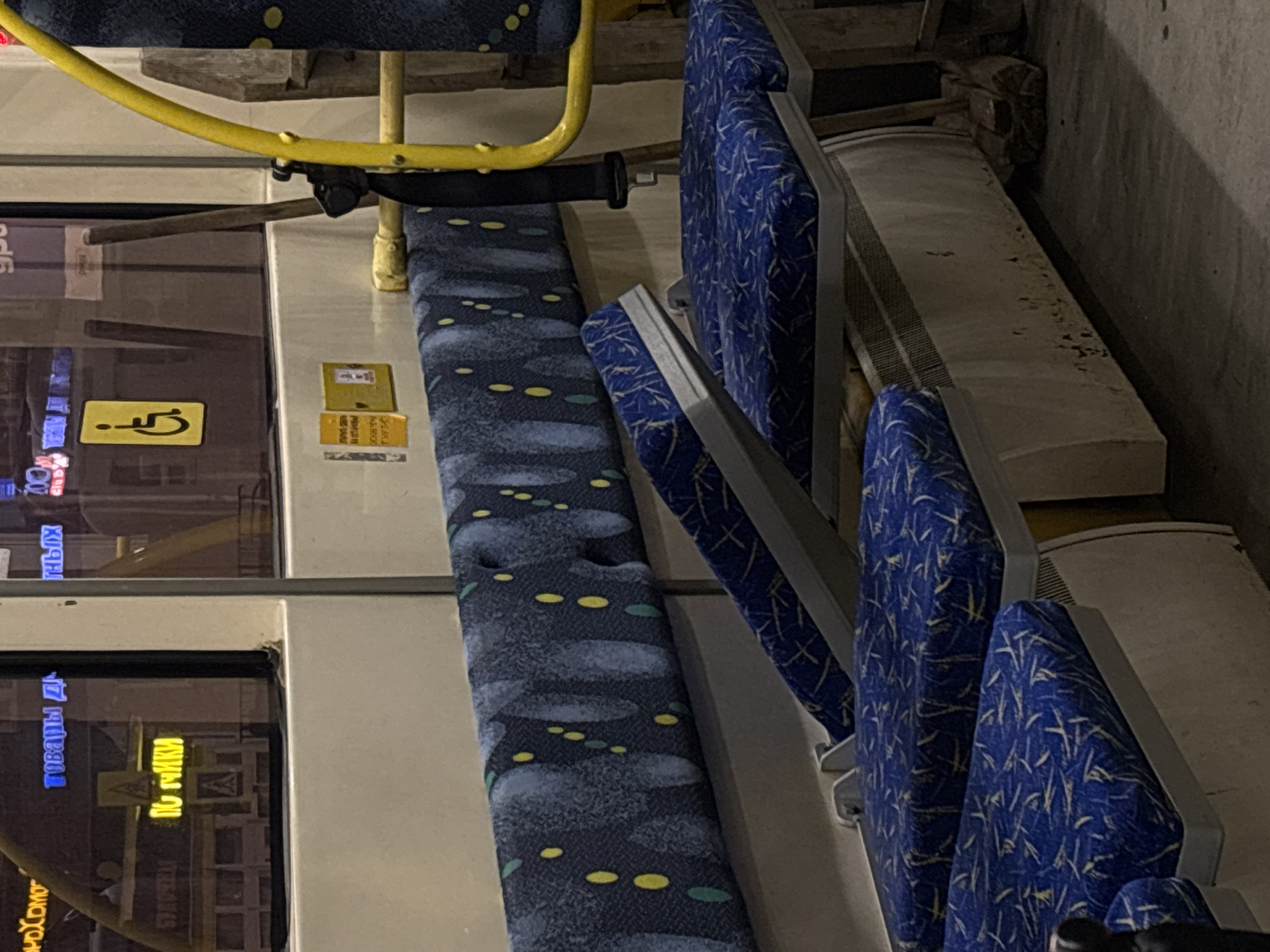 The image shows the interior of a bus, likely in Perm, Russia, based on the Cyrillic writing on the windows. There are rows of bus seats upholstered in blue patterned fabric. A yellow handrail runs above the aisle. A wheelchair accessibility sign is visible on the window. The area appears dimly lit.