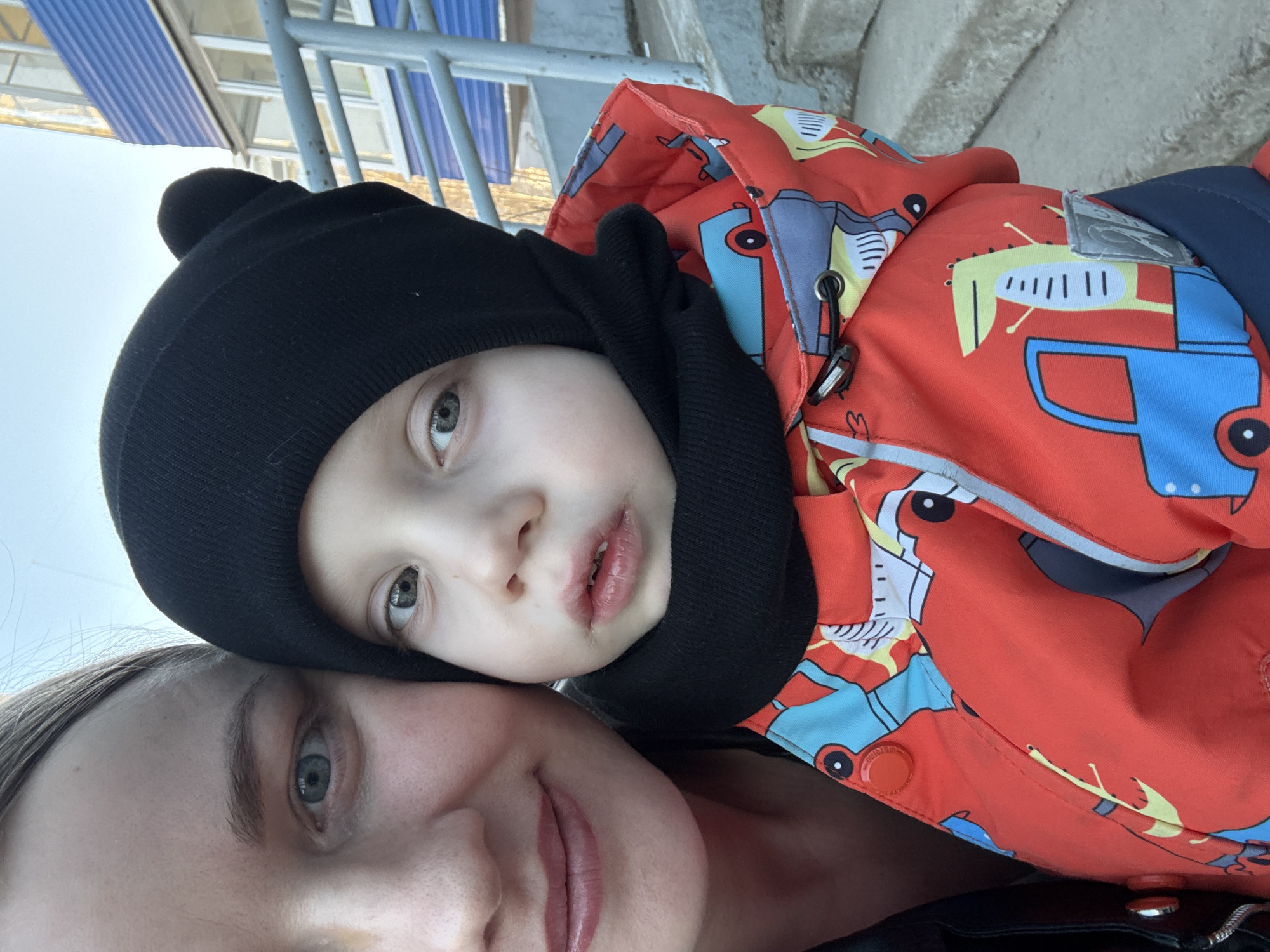 A close-up, tilted selfie shows a woman with a young child. The woman is at the bottom of the frame, her face partially visible, with light skin and light-colored eyes. The child is above her, wearing a black beanie and an orange jacket with cartoon car prints. The background is blurry but appears to be an urban scene, potentially in Chaikovsky, Russia, given the prompt. Elements like metal railings and a corrugated roof are visible, suggesting an outdoor setting possibly near a building. There are no visible interactions or actions occurring, the focus is on capturing the portrait.