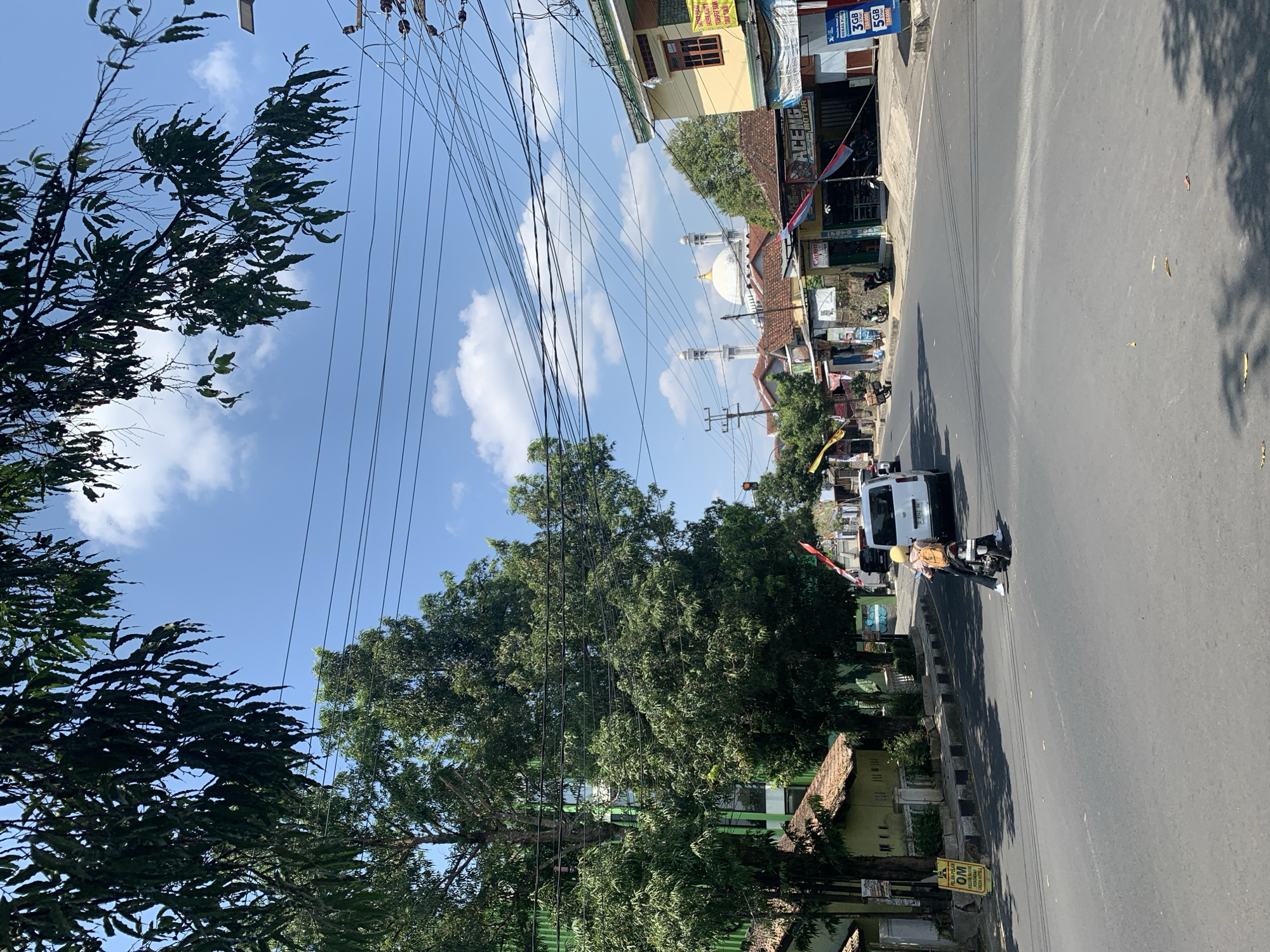 The image depicts a street scene in Ciamis, Indonesia, under a clear blue sky with scattered clouds. The foreground shows an asphalt road with a motorcycle carrying a person in a yellow hijab and an orange backpack, moving away from the camera. To the left of the road, a white van is visible, and beyond it, a building with a green facade and a brown tiled roof is present. Several trees with dense green foliage line the left side of the street, some with branches extending into the upper part of the frame.
The middle ground features a row of shops and buildings along the right side of the street. One shop has a yellow facade with a brown-trimmed window. Signs are visible on some buildings, including one with the text "ACENG MOTOR". A white mosque with two minarets and a prominent white dome is situated further down the street in the background, partially obscured by buildings and trees. Numerous electrical wires crisscross above the street and buildings, indicating urban infrastructure. The overall environment suggests a moderately busy street in a town or city, likely during daylight hours.