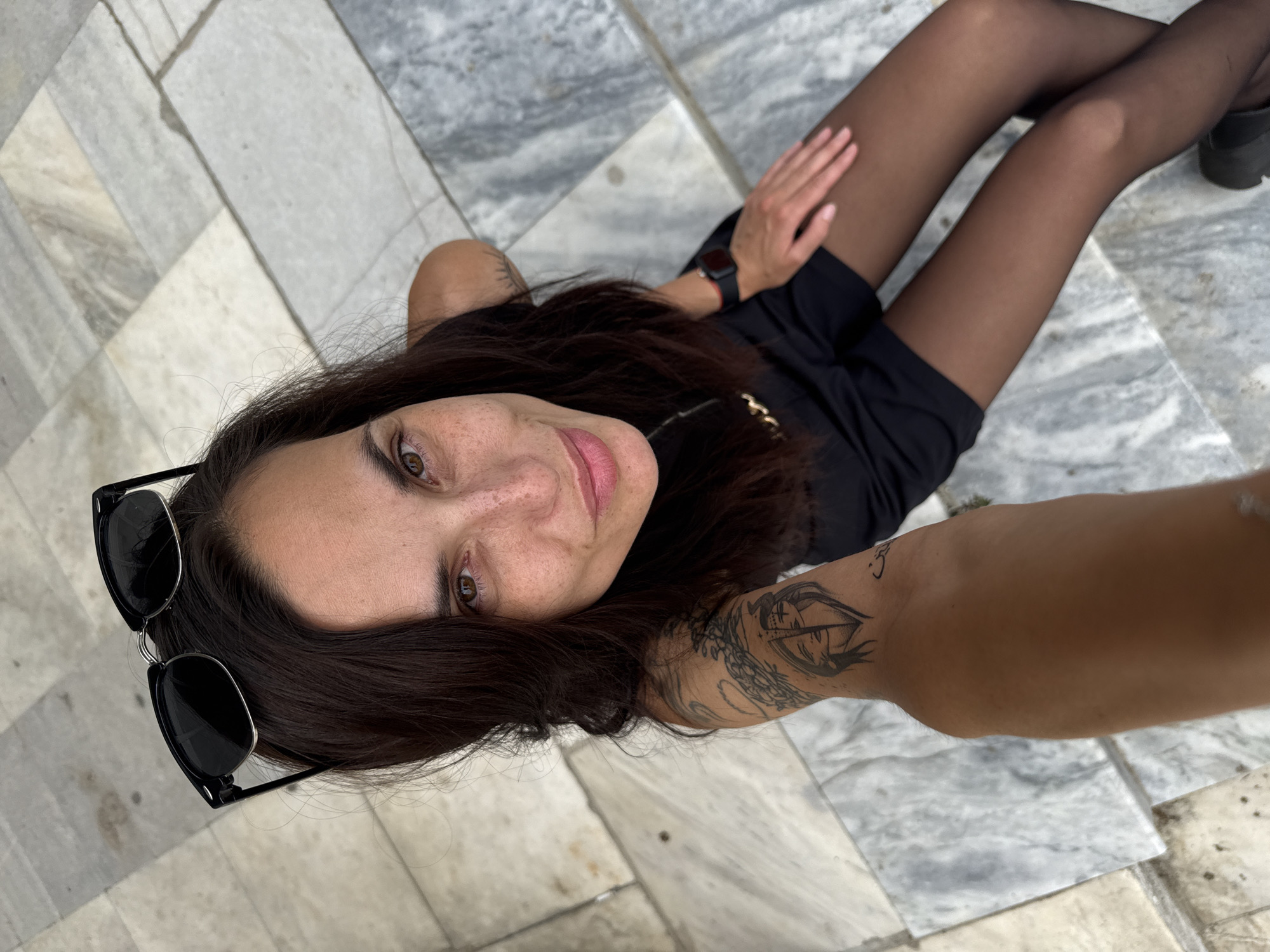A woman is seen from a high-angle perspective, sitting on a tiled surface. She has dark hair and is wearing a black dress with black tights. Sunglasses are placed near her head. She has visible tattoos on her arm. The tiled surface appears to be part of an outdoor public space, potentially in Mezdra, Bulgaria.