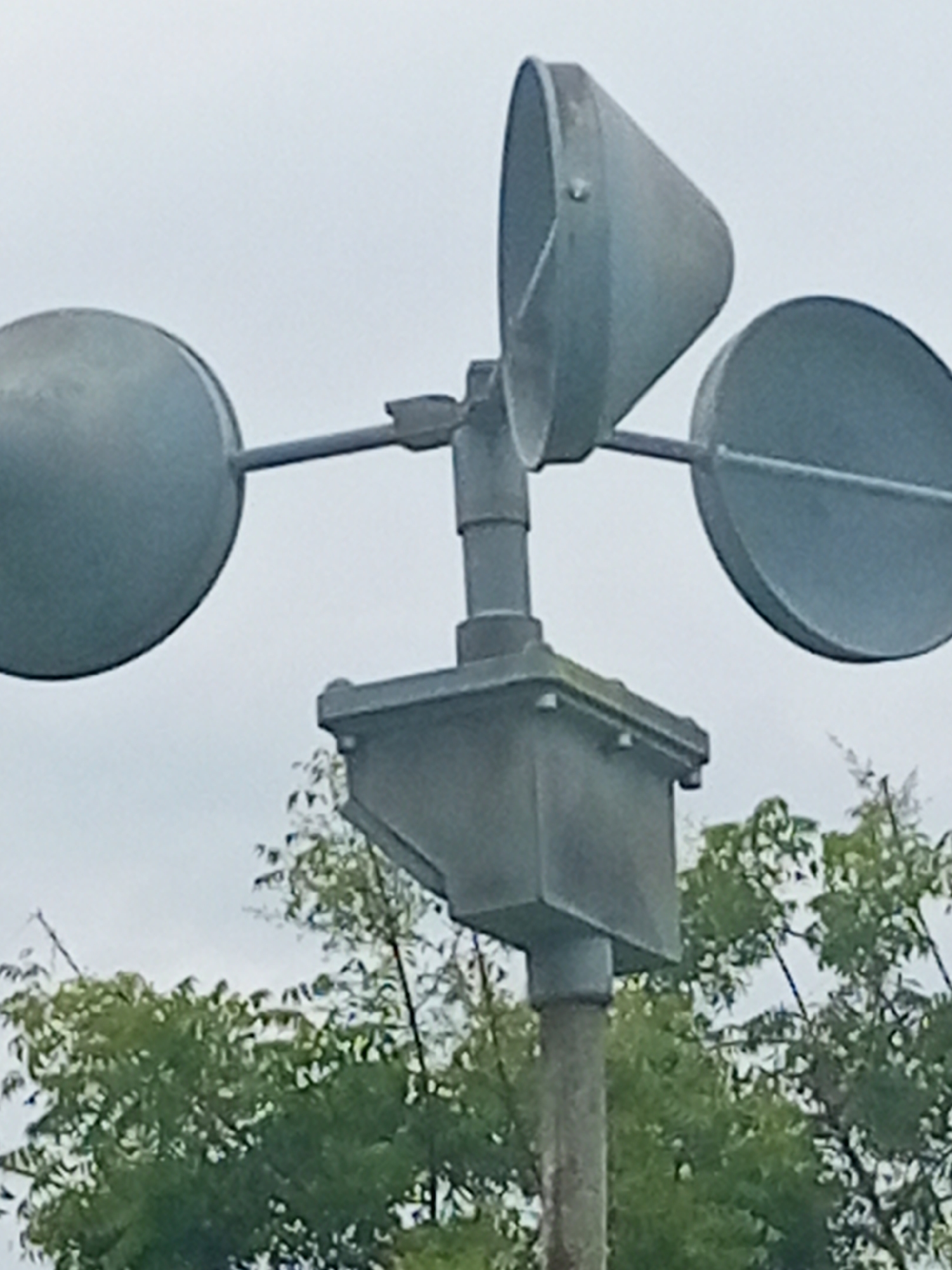 This is a close-up, medium-shot photograph of a weather vane, likely mounted on a pole. The weather vane consists of three cup-shaped elements attached to horizontal arms. Two of the cups are circular and positioned opposite each other, while the third cup is cone-shaped and positioned perpendicular to the other two. The weather vane is made of a dull, gray metal. Below the weather vane is a rectangular housing unit, also made of gray metal, attached to a central pole. The background is dominated by a cloudy, overcast sky and the tops of lush green trees. The overall impression is of a functional outdoor instrument. This object is likely part of a meteorological station or a weather monitoring device, and it is plausible that such equipment could be found in Jalingo, Nigeria, a city known for its climate research and agricultural activities.