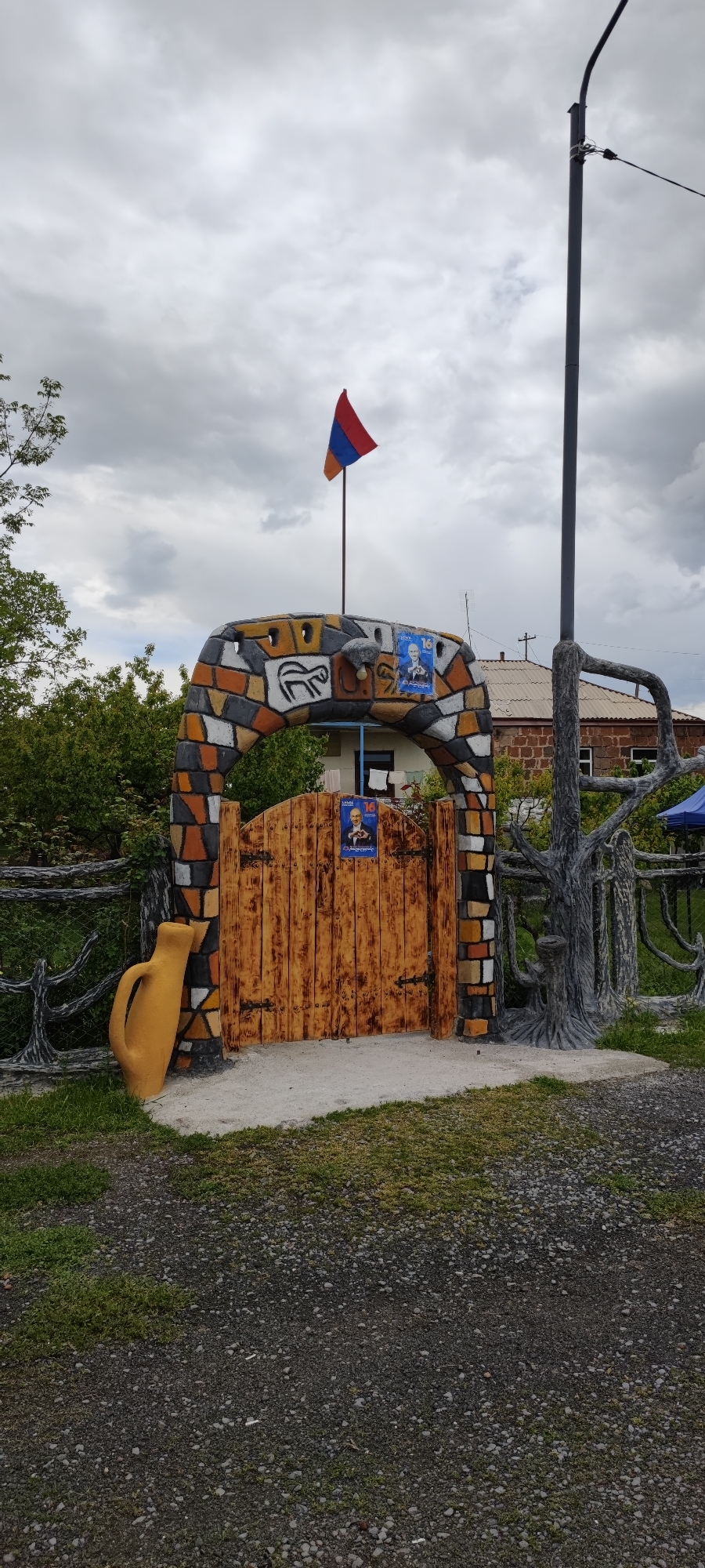 A wooden gate with a brick archway is situated in Dashtadem, Armenia. The archway is decorated with depictions of animals and portraits of a man. The Armenian flag is mounted on top of the archway. The gate is flanked by a yellow jar and a tree-like structure. The ground is covered with gravel and grass. A streetlight is visible in the background. A house can be seen through the gate. The sky is overcast.
