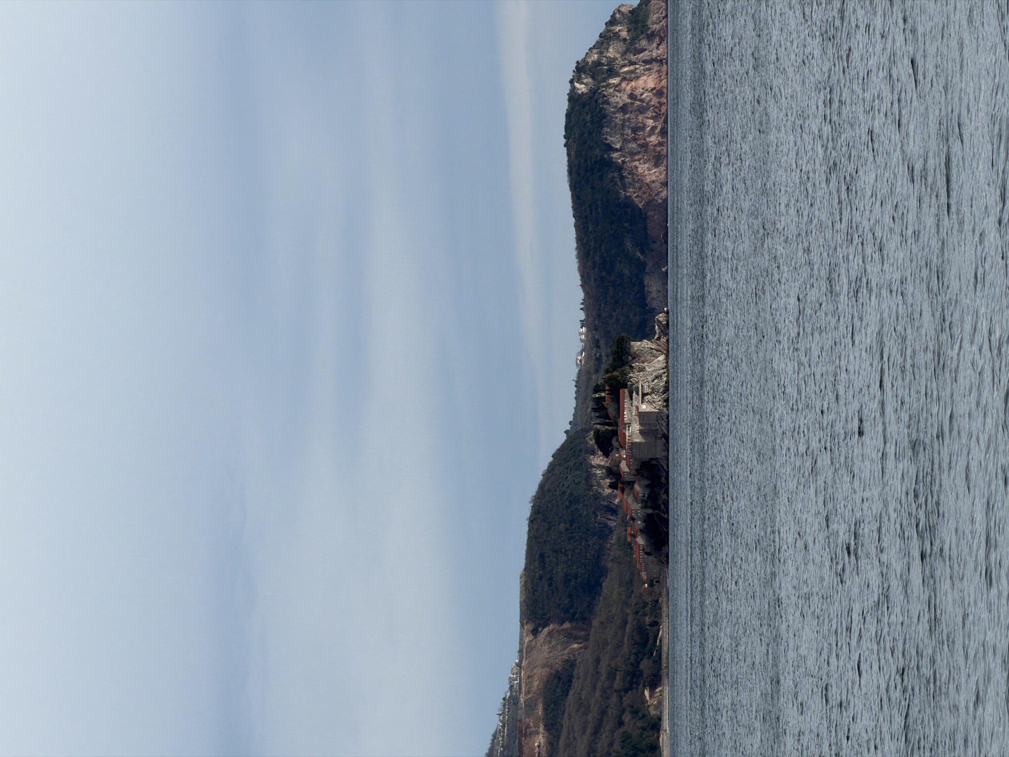 The image depicts a view of the coastline in Rafailovići, Montenegro. On the right is the Adriatic Sea, with visible surface texture due to light and waves. Adjacent to the sea is a mountainous terrain covered with vegetation. Buildings, including Sveti Stefan, are visible on the cliffs. The sky above is light blue with faint cloud formations. The overall composition is horizontally oriented.