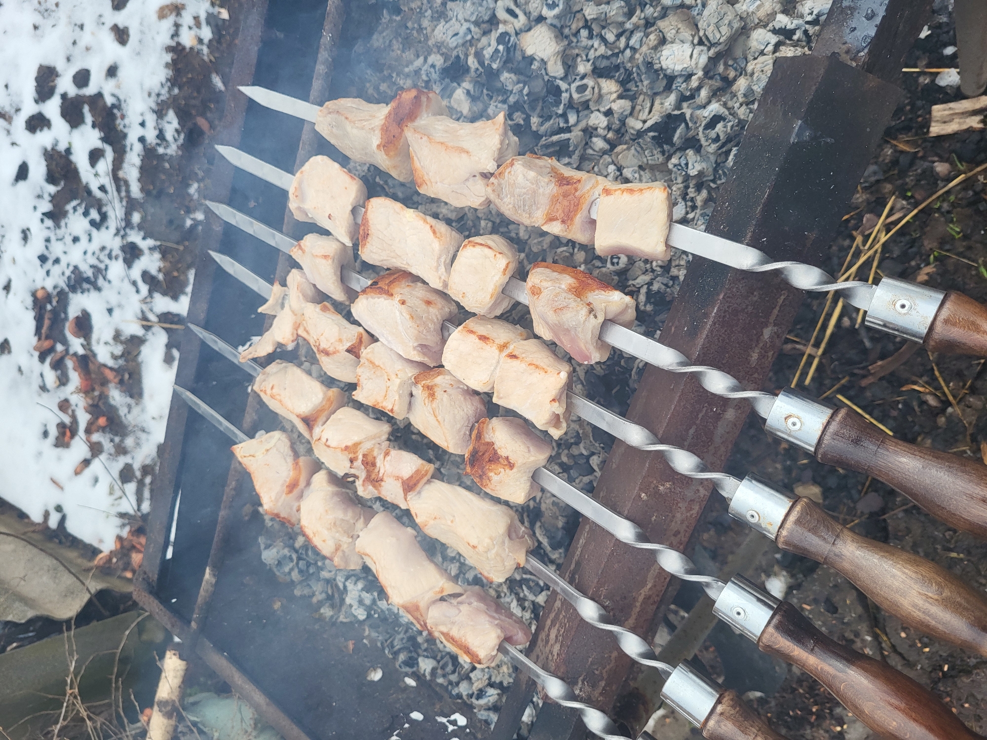 The image shows four skewers of meat, possibly pork or chicken, cooking over a charcoal grill. The skewers are made of metal with twisted sections and wooden handles. The meat is cut into roughly cube-shaped pieces, some with visible browning. The grill sits outdoors, with some snow visible in the background, along with foliage. The location is Зипуново, Russia.