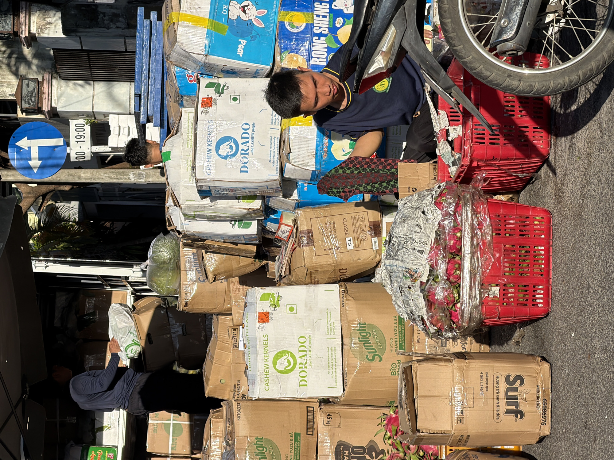 A street vendor in Nha Trang, Vietnam, is surrounded by stacked boxes of goods, including cashews, and a red basket filled with pink dragon fruit. The vendor, a young man in a dark blue shirt with yellow trim, is seated and appears to be organizing or handling some items. The scene is outdoors, with a white awning overhead and a sidewalk visible. A motorcycle is parked nearby. The overall impression is of a busy market or roadside stall.