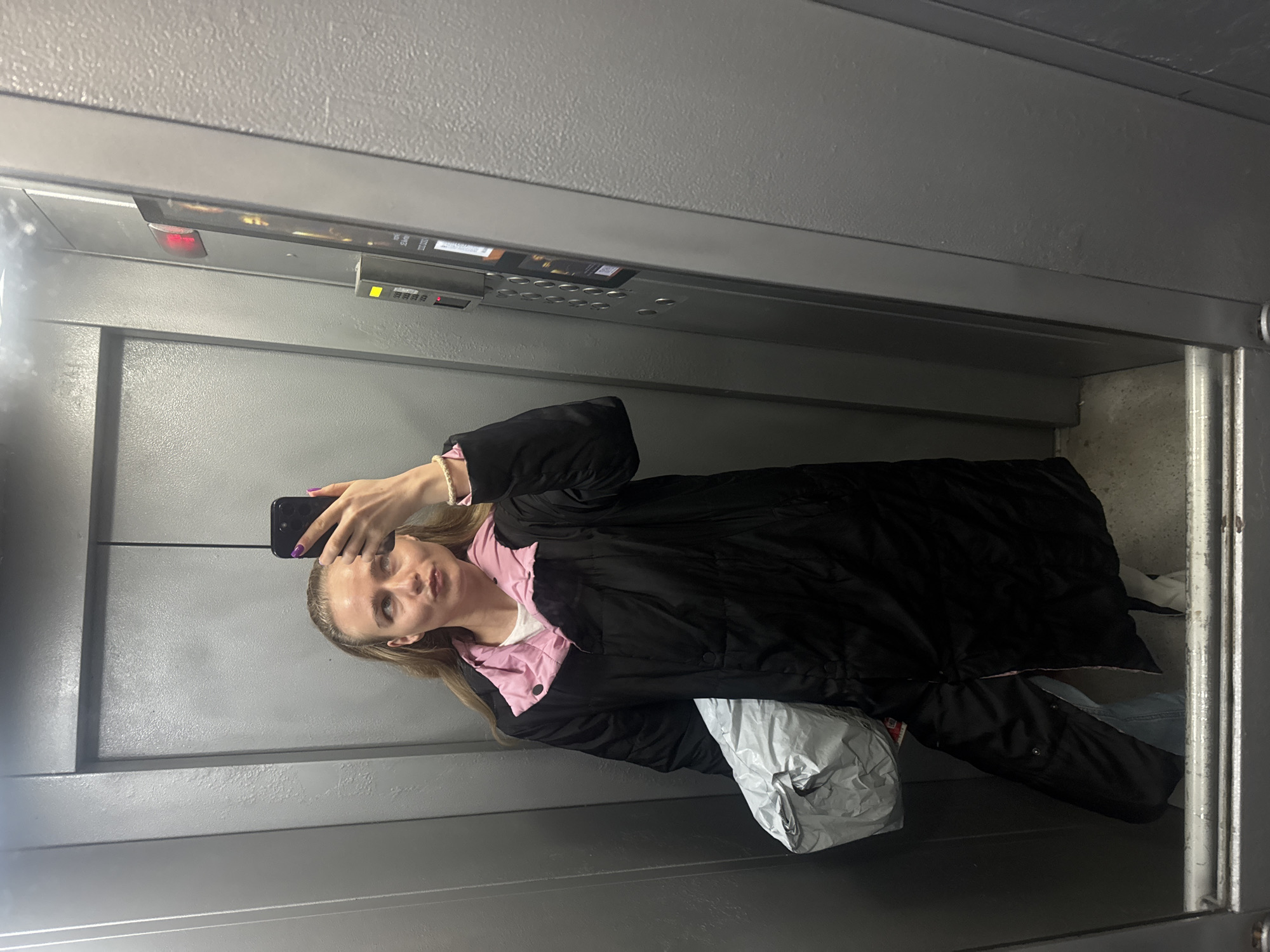 A woman takes a selfie in an elevator. She is wearing a black coat with a pink collar and is holding a black smartphone in her right hand. She is also holding a silver package. The elevator has gray walls and a control panel at the top. The location is Unknown, Unknown.