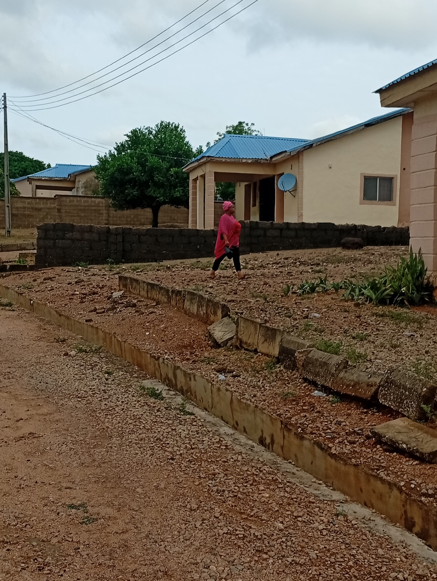 A woman in a pink headscarf and top walks across a gravel-covered area adjacent to a concrete drainage ditch in Adankolo, Nigeria. Behind her is a low stone wall and a tan-colored house with a blue roof and satellite dish. Another similar house is visible in the background, along with a tree. Power lines run overhead. The sky is overcast.