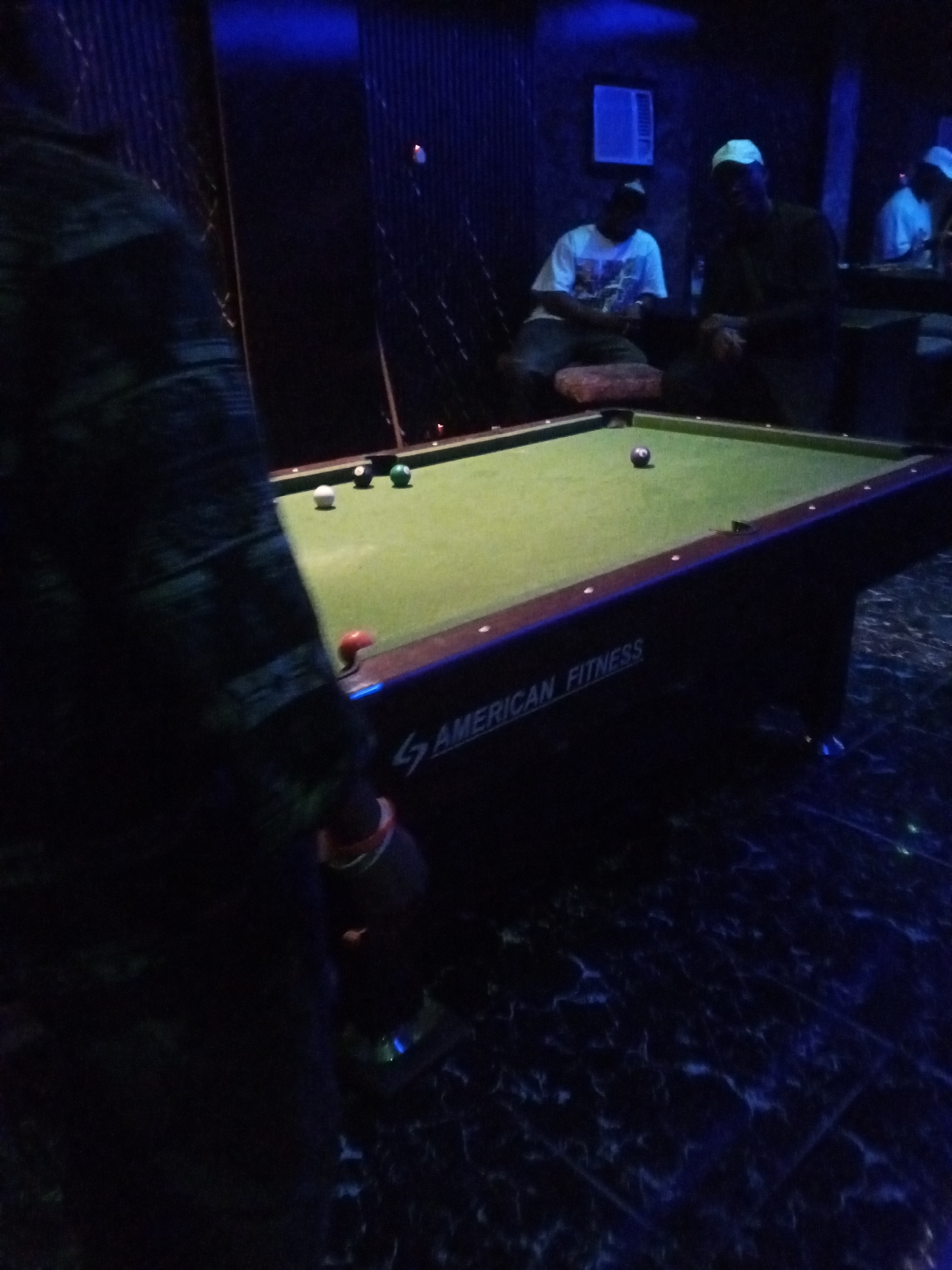 In a dimly lit establishment in Lokoja, Nigeria, a pool table with a green felt surface is the central focus. Several billiard balls are scattered across the table, including a black ball near the center. Two men, one wearing a white t-shirt and the other a dark shirt and a light-colored cap, are seated on the floor near the table. A third man, partially visible on the left side of the frame and facing away from the camera, is wearing a dark, patterned shirt. The room is decorated with dark, ribbed paneling on the walls and features blue lighting, with an air conditioning unit visible on the far wall. The floor has a dark, marbled pattern. The overall atmosphere suggests a casual social gathering centered around playing pool.