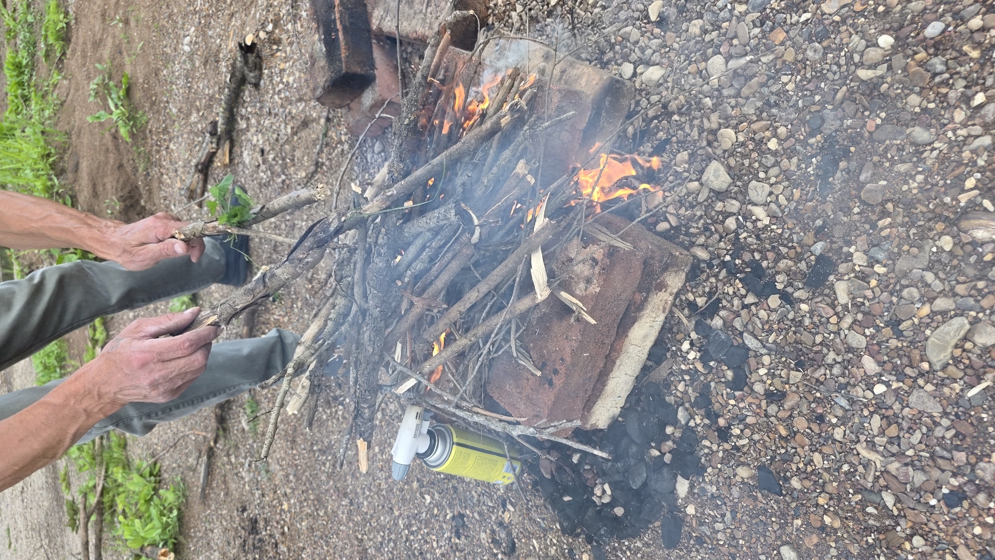 In Дарьино, Russia, a person's hands are adding dry twigs to a small bonfire. The fire is situated on a patch of gravel, with a few pieces of brick serving as a makeshift fire pit. A yellow butane torch is placed beside the fire pit. The background shows dry soil and sparse green vegetation. The overall scene depicts the process of starting an outdoor fire.