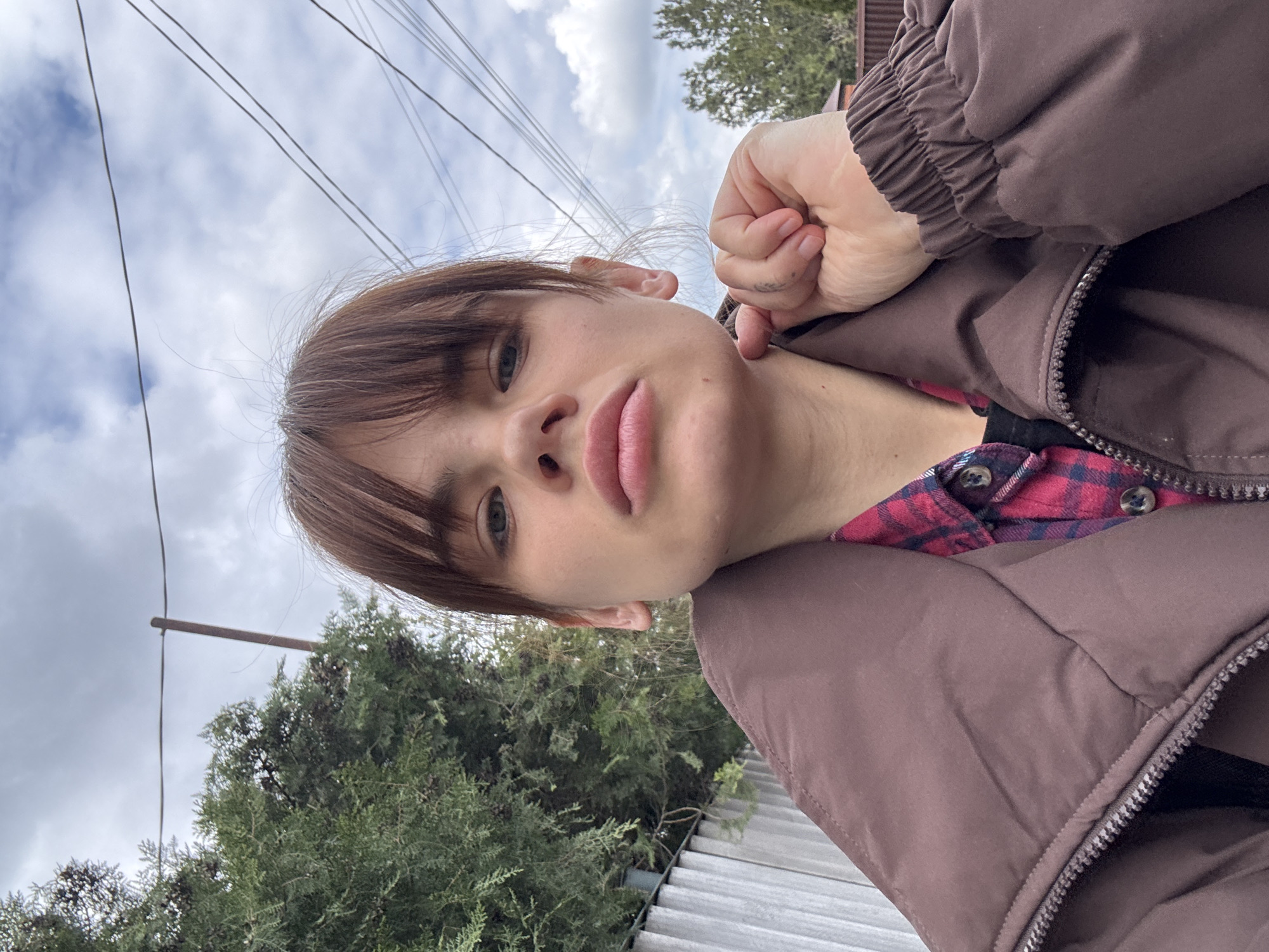 A selfie taken outdoors depicts a young person with fair skin, brown hair with bangs, and green eyes. The person is wearing a brown puffer jacket over a red and black plaid shirt. Their hand is near their face, touching their jawline. The background shows trees, a fence, and a cloudy sky with power lines. While the image provides visual details, it doesn't contain explicit information to confirm the location as Rostov-on-Don, Russia. This would require additional information outside the image context.