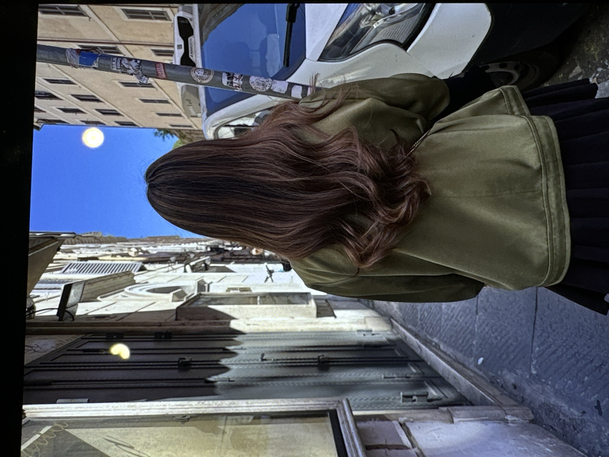 The image shows a woman with long, wavy brown hair wearing an olive green jacket and a black skirt. She is standing on a city street with buildings on one side and a car parked on the other. The buildings appear to be old and made of stone, with multiple windows. There is a blue sky visible in the background, indicating it is daytime. The image is rotated.
While Yerevan, Armenia is a possible location, confirmation would require more contextual details from the photograph, such as distinct architectural elements or signage unique to Yerevan.