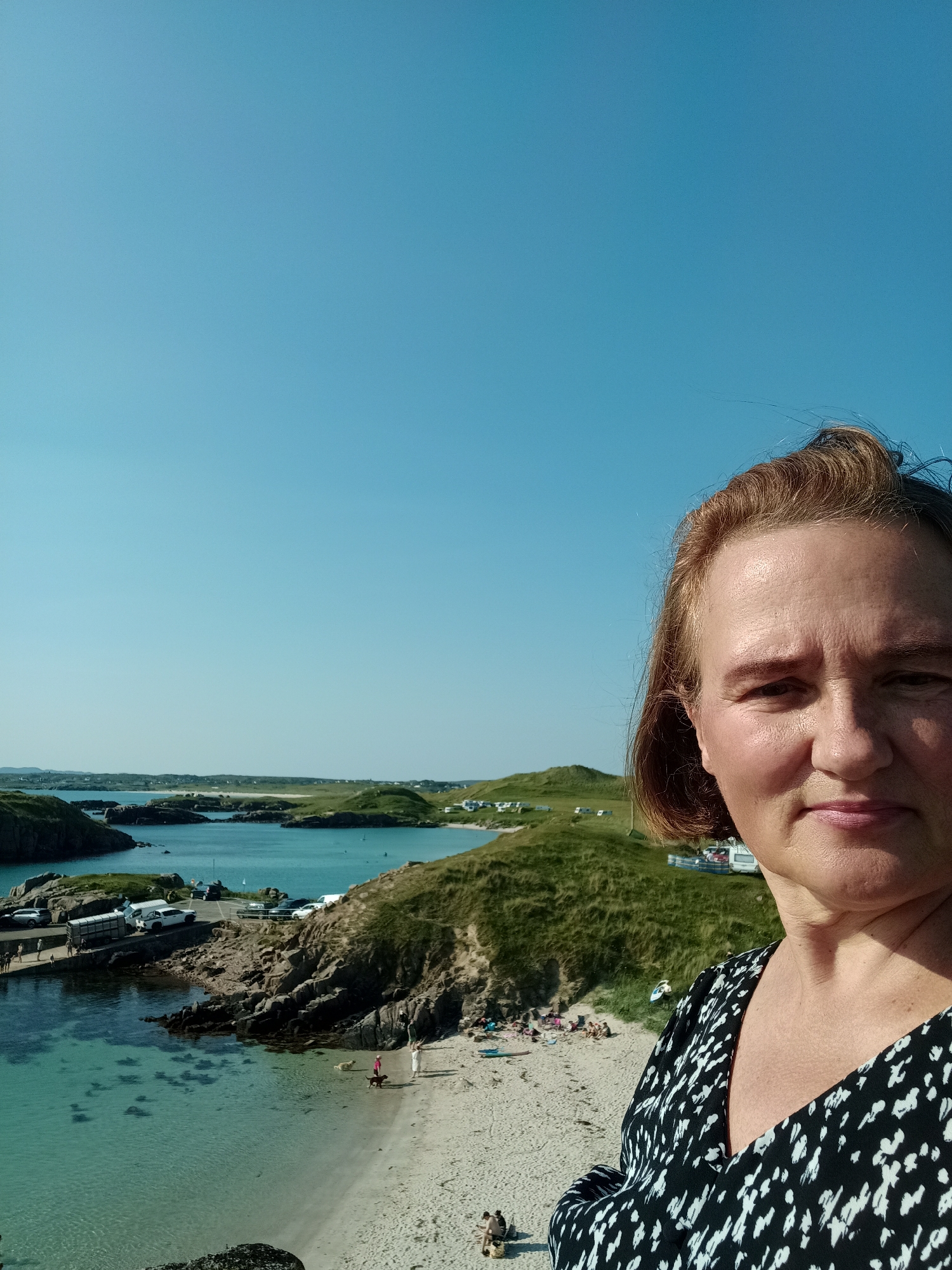 A woman, facing the camera, stands in the foreground of this image, her upper body and face dominating the right side. She has auburn hair and is wearing a black and white patterned dress. Behind her, a picturesque coastal landscape unfolds under a clear blue sky. The scene is the town of Carnboy, Ireland. A small bay with crystal-clear turquoise water is visible, with a sandy beach dotted with a few people. Rocky outcrops and green, grassy hills form the coastline. In the bay, several boats are anchored, and a small pier is visible with cars and a trailer parked nearby. In the background, more green hills rise, and some buildings and what appear to be mobile homes or caravans are scattered across the landscape. The overall impression is of a sunny, idyllic day at the seaside.