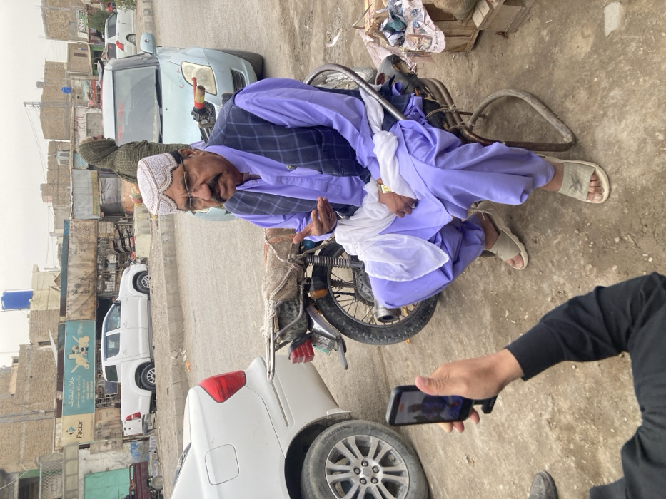 The image depicts an outdoor street scene in Taftan, Pakistan, under an overcast sky. In the foreground, the lower torso and arms of an individual wearing a black long-sleeved shirt are visible, holding a black smartphone vertically. The phone's screen displays a live view or recorded image of the primary subject. The primary subject is an adult male with a mustache and eyeglasses, dressed in a light purple shalwar kameez, a dark blue checkered vest, and a white, intricately patterned traditional cap. He is seated on a simple metal chair, wearing light-colored open-toed sandals. A white cloth is draped over his lap, and a gold-colored watch is visible on his left wrist. His right hand is raised and open, performing a gesture indicative of speech or explanation.
Directly behind the seated man and to his left is a parked dark-colored motorcycle featuring chrome components and a fabric-covered seat. Further back, a light blue compact car is parked. Behind this vehicle, a person with a patterned head covering is partially visible. On the far left of the frame, the rear section of a white sedan is visible.
The background consists of a street with unpaved and dusty ground areas, alongside sections of asphalt. Buildings line the street, appearing as single-story or two-story structures made of light-colored masonry, some with exposed construction elements. Prominent signage on one building includes the word "Factor" and the Urdu text "حلال الیکٹرک" (Halal Electric). A blue water tank is visible on the rooftop of one of the buildings in the mid-background. A white pickup truck is parked on an elevated, unpaved section of the street further back.