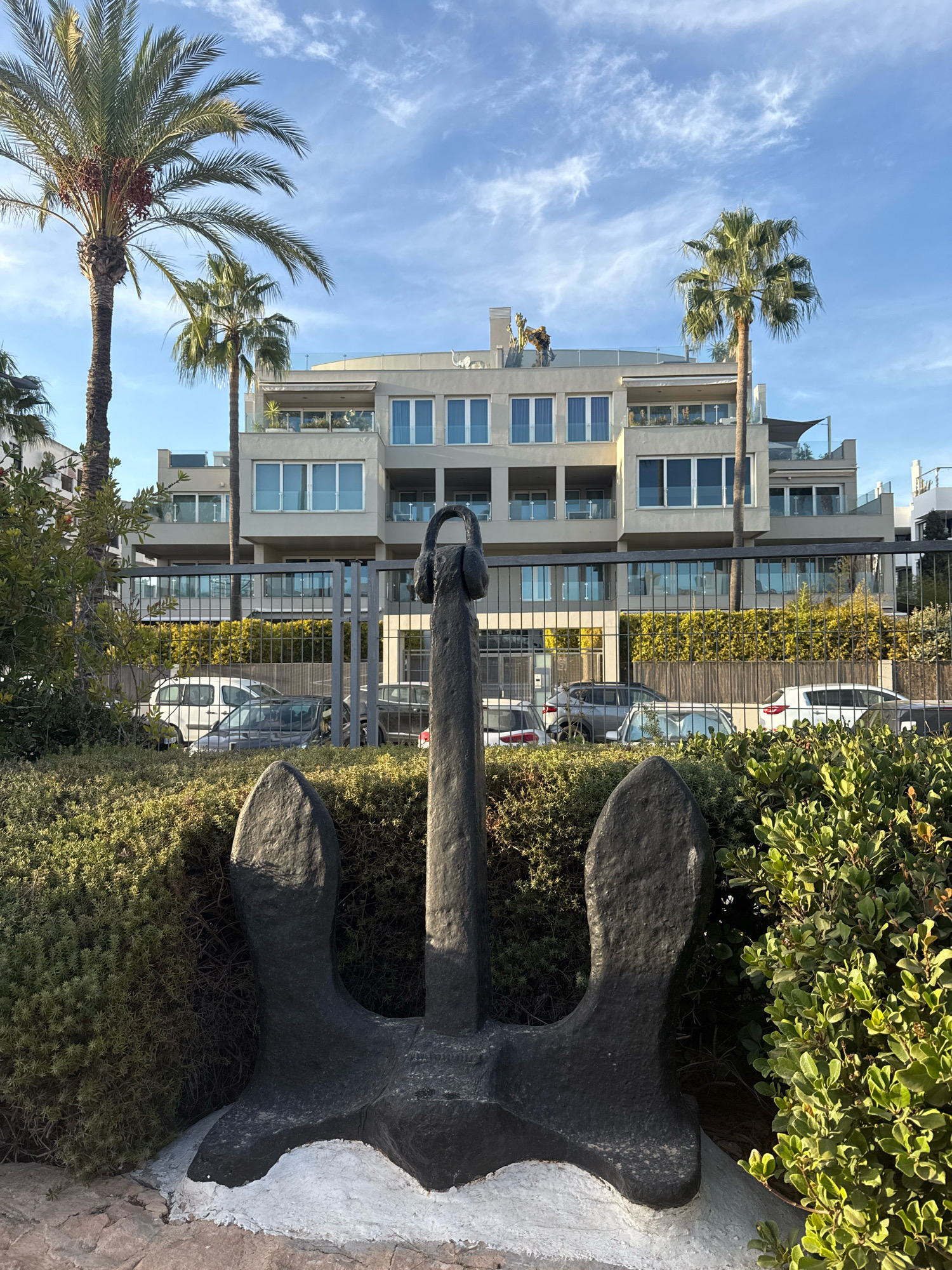 In Ibiza, Spain, a large, dark anchor sculpture with what appears to be headphones on top stands in the foreground, partially obscured by lush green bushes. Behind the anchor, a black metal fence separates the viewer from a street where several cars are parked. Beyond the fence and cars, a modern, multi-story building with many windows and balconies is visible. Tall palm trees flank the building, and the sky above is blue with scattered clouds. The scene is bathed in sunlight, casting shadows and highlighting the textures of the anchor, foliage, and building.