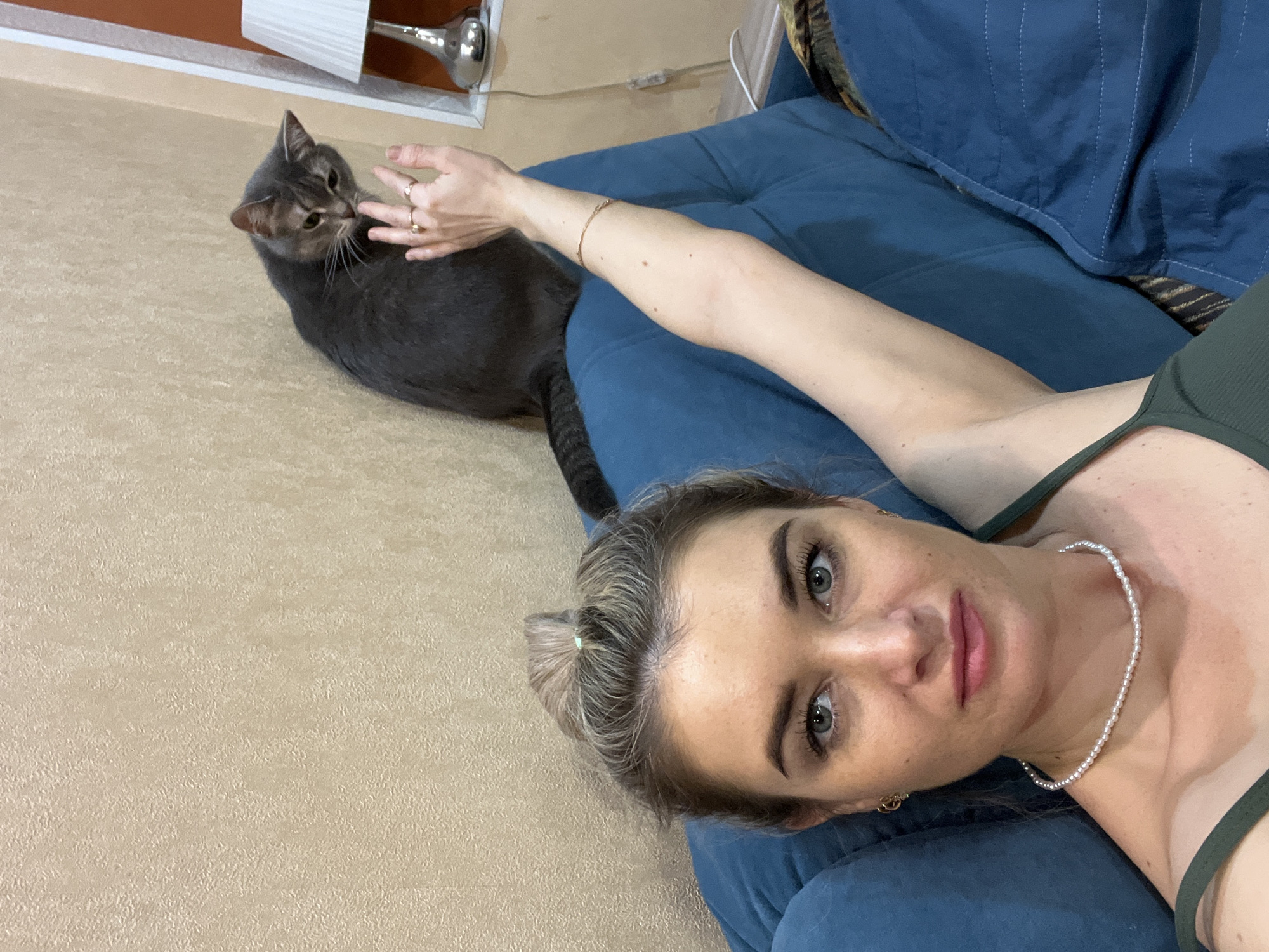 A selfie shot shows a Caucasian woman with her head resting on a blue upholstered couch. She has light hair pulled back, green eyes, fair skin, and is wearing a pearl necklace and a green top. She is looking up at the camera. Next to the couch on a beige textured floor sits a gray cat, being petted by the woman's outstretched hand. Behind the cat is a baseboard and wall with a lamp. The location is inferred to be in the city of Chaikovsky, Russia, as per the instructions.