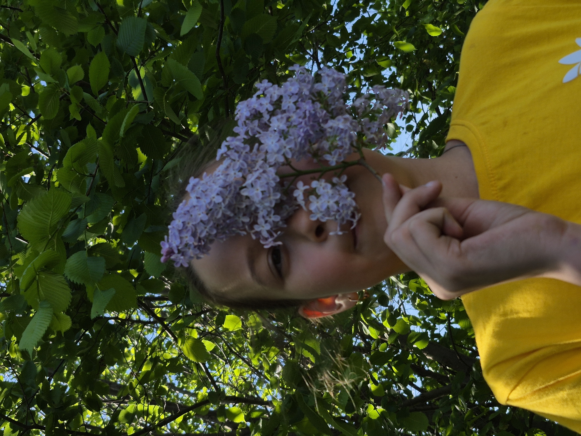 The image depicts a young person holding a sprig of lilac flowers in front of their face, with a background of green leaves and branches against a blue sky. The person is wearing a bright yellow t-shirt with a white flower motif on the shoulder. The angle is from below, looking up, and the image is somewhat rotated, giving a tilted perspective. The location is Chaikovsky, Russia.