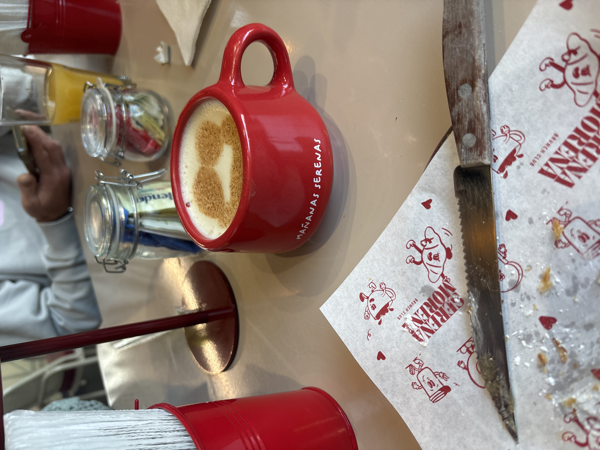 The image depicts a tabletop scene, likely inside a café in Puebla, Mexico, evidenced by the "Mañanas Serenas" branded red mug filled with a coffee drink, and the "Serena Morena Brunch Club" branded paper napkins. A serrated knife rests atop one of the napkins, which shows crumbs or leftover food particles. Near the mug, there are glass jars containing condiments or sugars. In the background, a partial view of a person's hand and a red metal container holding white napkins are visible. The table surface is a light tan color.