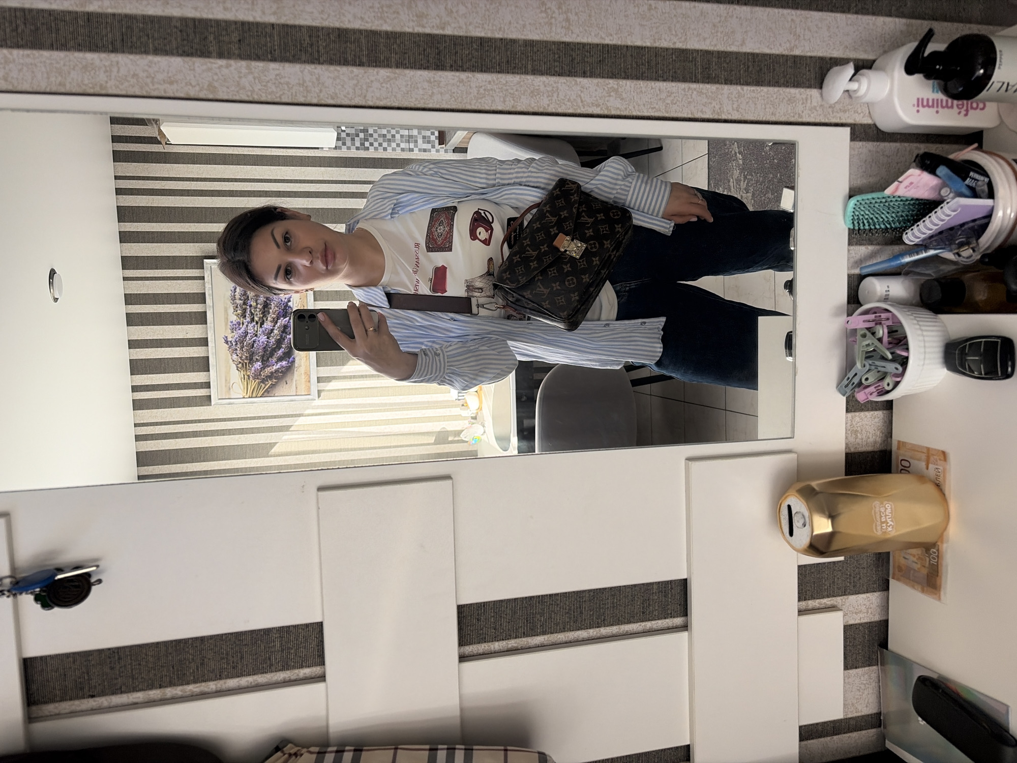The original image is rotated 90 degrees counter-clockwise, showing a woman taking a mirror selfie in an indoor setting.
The woman, reflected in a large, white-framed mirror, is captured from the chest to the mid-thighs. She has dark hair pulled back and a neutral expression. She wears a white t-shirt with graphic prints of fashion accessories, layered under an unbuttoned light blue and white striped long-sleeved shirt, and dark blue jeans. A dark brown Louis Vuitton monogrammed cross-body bag with a gold clasp hangs across her body. She holds a black smartphone, likely an iPhone, in her right hand, capturing her reflection. A ring is visible on her right hand.
The background behind the woman, visible in the mirror and around it, features a light gray and white striped wallpaper. Within the reflection, a framed picture depicting lavender flowers is seen on the wall, with a notable shadow suggesting natural light.
On a white surface to the right of the mirror, various personal items are arranged. These include a white pump bottle labeled "café mimi" and "VANILLE...", a white ceramic holder containing hairbrushes (one turquoise), notebooks, and pens, and another white pot with light blue and pink clothes pins. A black car key fob is also present. A gold-colored, can-shaped object, possibly a piggy bank, displays Cyrillic text: "УЛЫБАЙСЯ" (Smile) and "И ВСЁ БУДЕТ КУПЛЮ" (And everything will be bought). Several Russian banknotes are visible underneath it, one clearly marked "100 РУБЛЕЙ" (100 Rubles). To the left of the mirror, a set of keys with a blue charm hangs on a hook. The scene suggests a casual, lived-in personal space during the daytime.