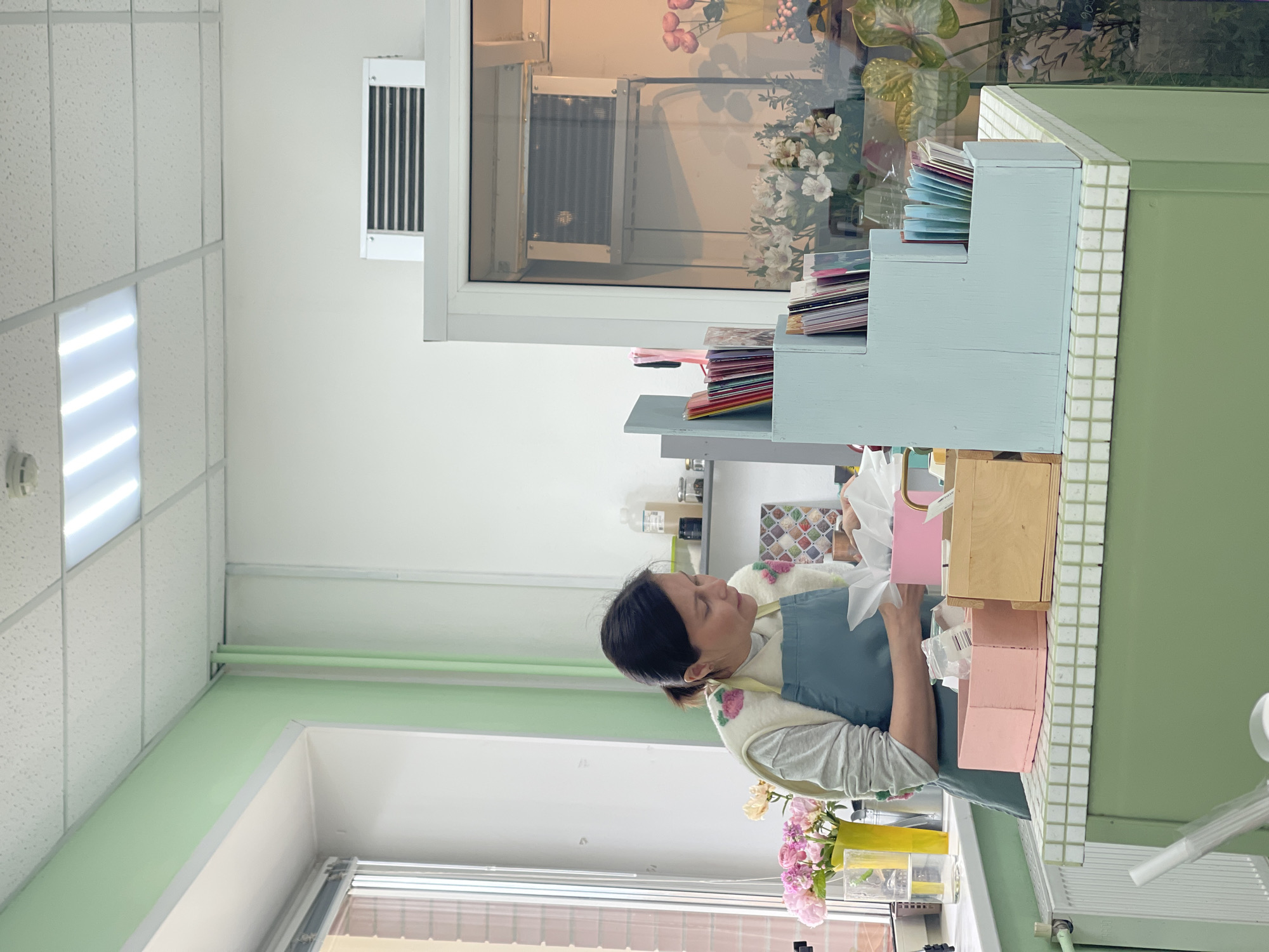 A woman is depicted actively working at a counter in what appears to be a flower shop, gift shop, or crafting studio. She has dark hair tied back and is wearing a light-colored long-sleeved shirt, a cream-colored vest adorned with floral embroidery, and a teal apron. Her focus is on the task at hand, involving white tissue paper and several pink boxes/containers on the counter directly in front of her.
The counter features a tiered light blue wooden organizer filled with colorful cards or papers, a simple wooden box, and a small vase holding pink flowers, possibly roses. Behind the woman, a large display window reveals various flowers and greenery, suggesting a floral display or refrigerator. Above this window, a white air vent is installed on a white wall. The ceiling consists of white acoustic panels with bright fluorescent strip lighting. The walls are painted in clean white and a soft pastel green. Another window is visible below the counter level, allowing natural light into the space. The scene is well-lit, indicating it is daytime. The overall ambiance is clean, organized, and focused on creative work. No discernible text is clearly visible on any objects or surfaces within the image.