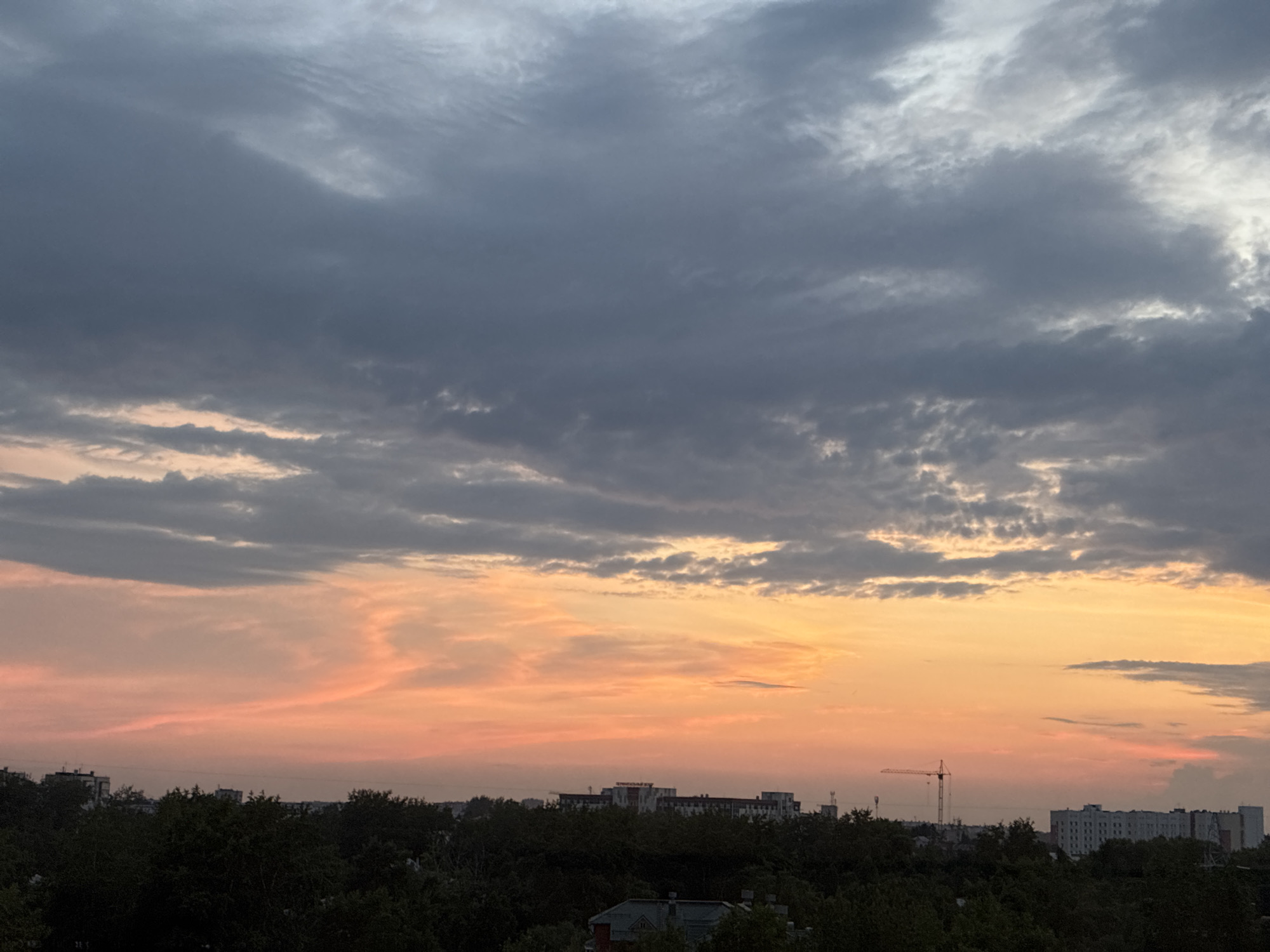 The image captures a dusky sky over a city skyline. The upper portion of the sky is filled with heavy, dark gray clouds. Below these, the clouds transition to softer, lighter gray, and then to a vibrant band of orange and pink as the sun sets. Silhouetted against this colorful sky is a dense line of dark green trees, interspersed with various buildings of different heights and architectural styles. Some buildings appear to be residential blocks, while others are more industrial or commercial, with visible windows and roofs. A tall construction crane is prominently visible in the middle ground, standing out against the sunset. The overall impression is one of a city transitioning from day to night, with the dramatic colors of the sunset painting the sky.