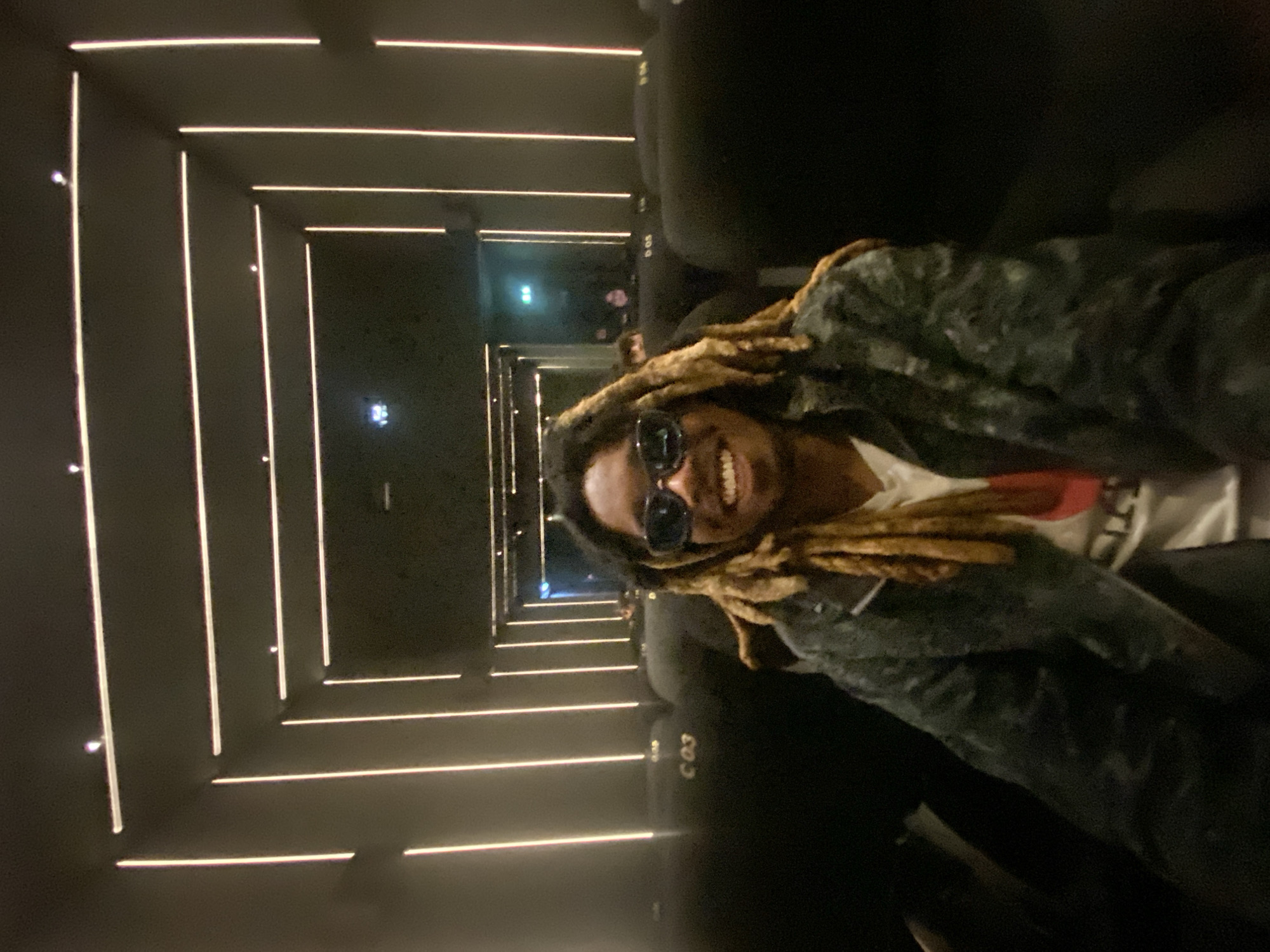 A person with dreadlocks and sunglasses is smiling in the seat of a movie theater. The theater's walls are lined with horizontal stripes of light. The row of seats where the person is sitting is labeled 'C 03'. The setting could plausibly be a cinema in Paris, France, due to its modern design, but this cannot be confirmed definitively from the image alone.